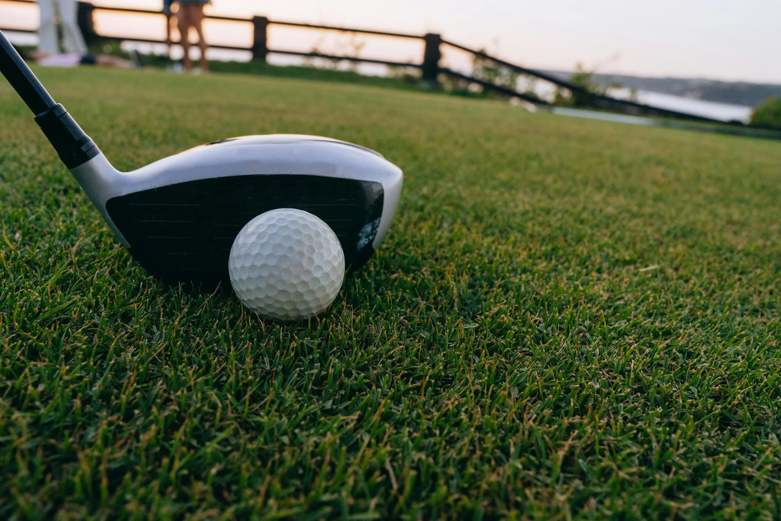 Close-up of a golf club and a golf ball on a grassy golf course during sunset with a distant view of a body of water and a wooden fence.