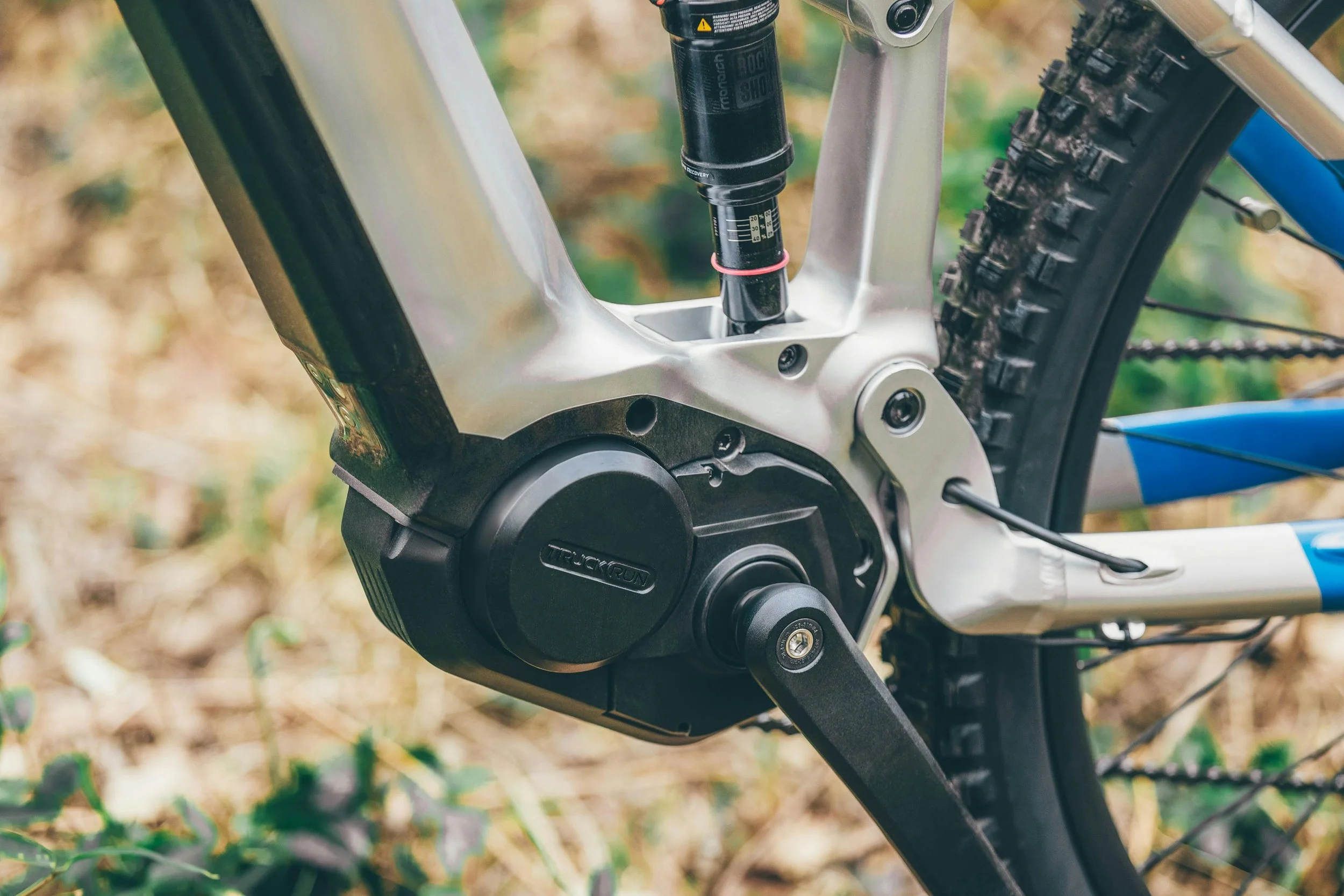Close-up of an electric mountain bike's motor and rear suspension system with a knobby tire in the background.
