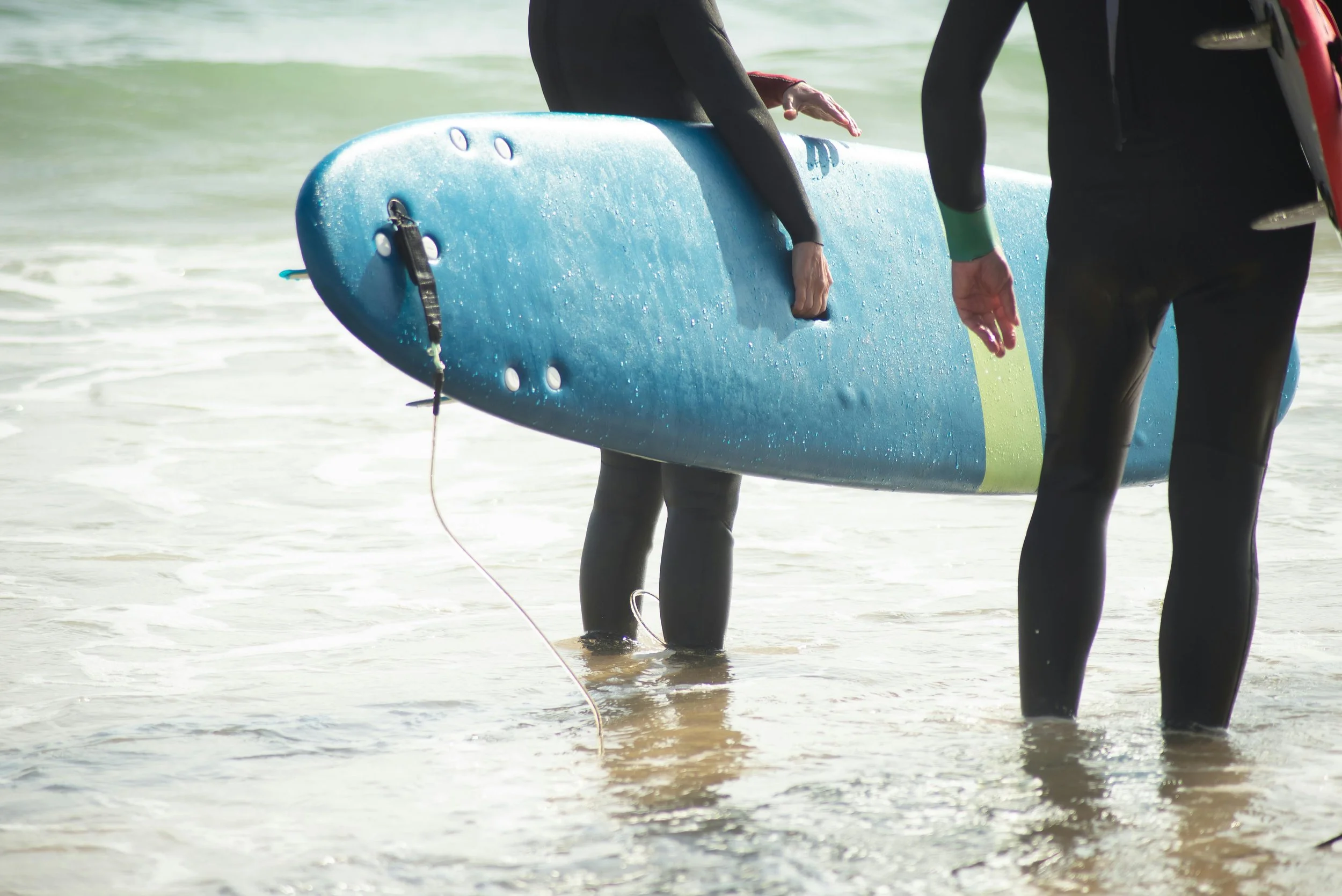 Two people in wetsuits standing in shallow water, holding a blue surfboard.