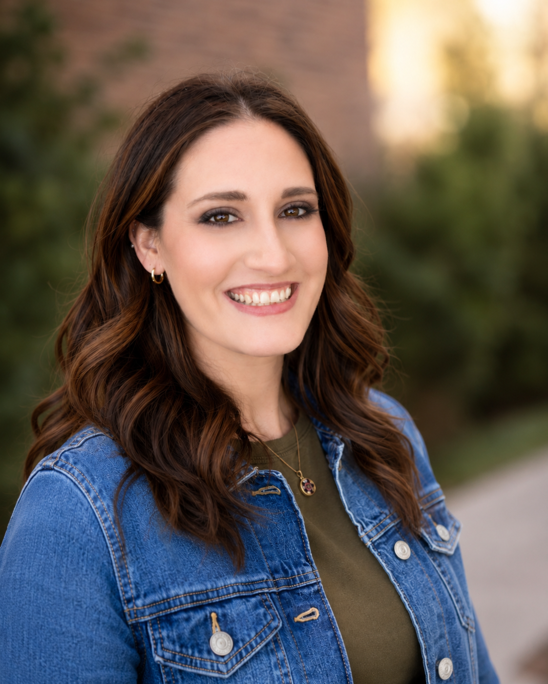 A woman with long wavy brown hair, smiling, wearing a denim jacket over an olive green top, standing outdoors with blurred greenery in the background.