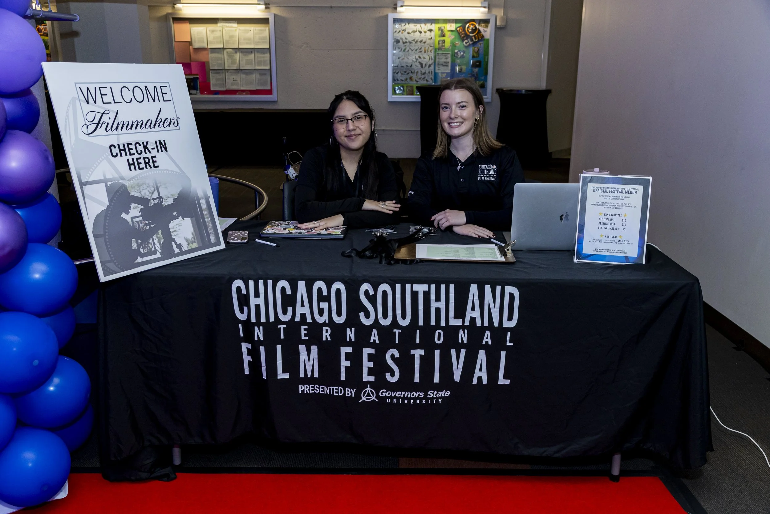 Two women sitting at a registration desk with a sign that reads "Welcome Filmmakers, Check-In Here" at the Chicago Southland International Film Festival, with informational posters on the wall behind them, and blue balloons on the left side of the fr