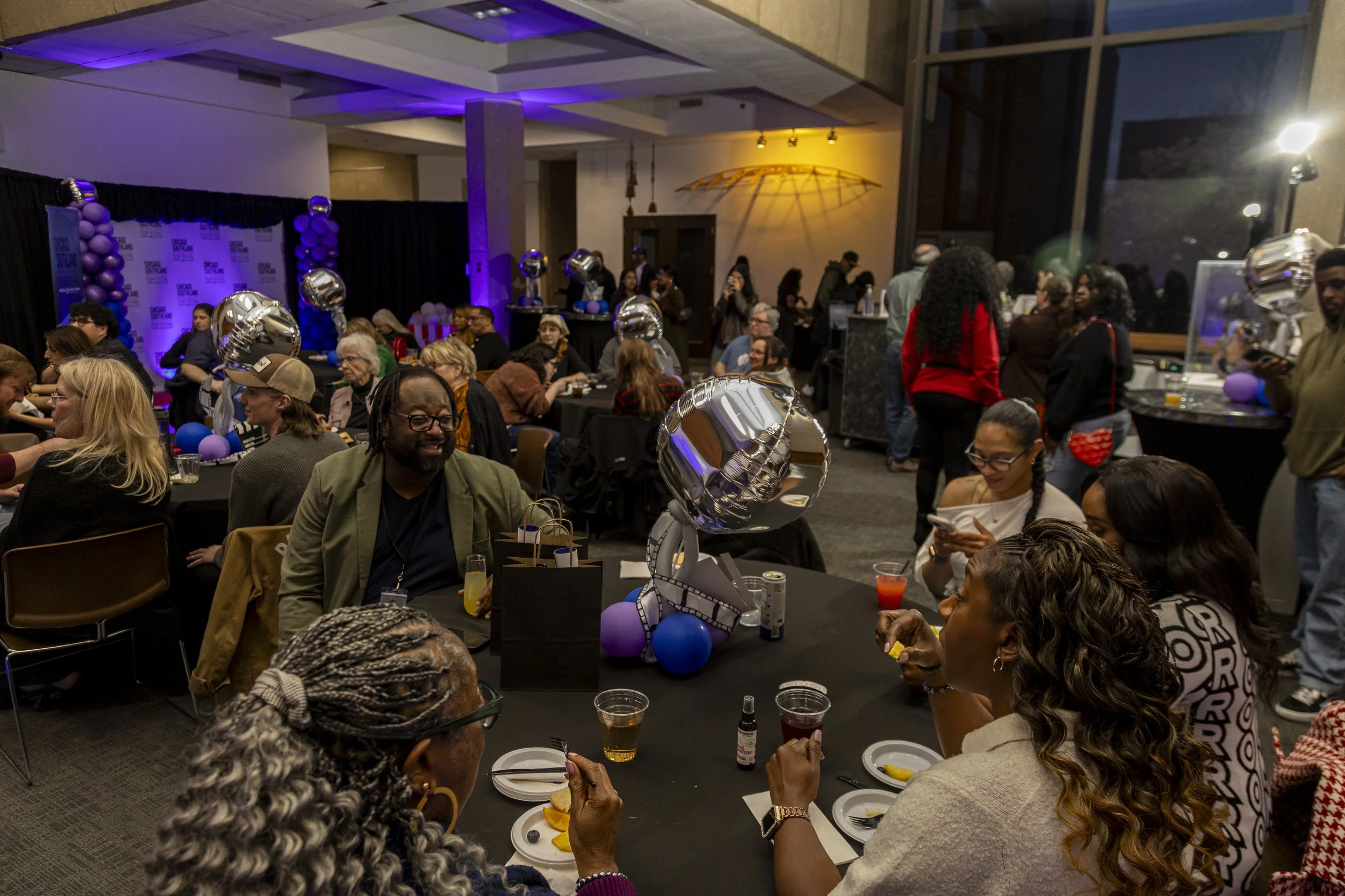 A large gathering of diverse people at a celebration or party indoors, decorated with balloons, including silver foil balloons and purple balloons, with some people sitting at tables and others standing or walking around, there are drinks on the tabl
