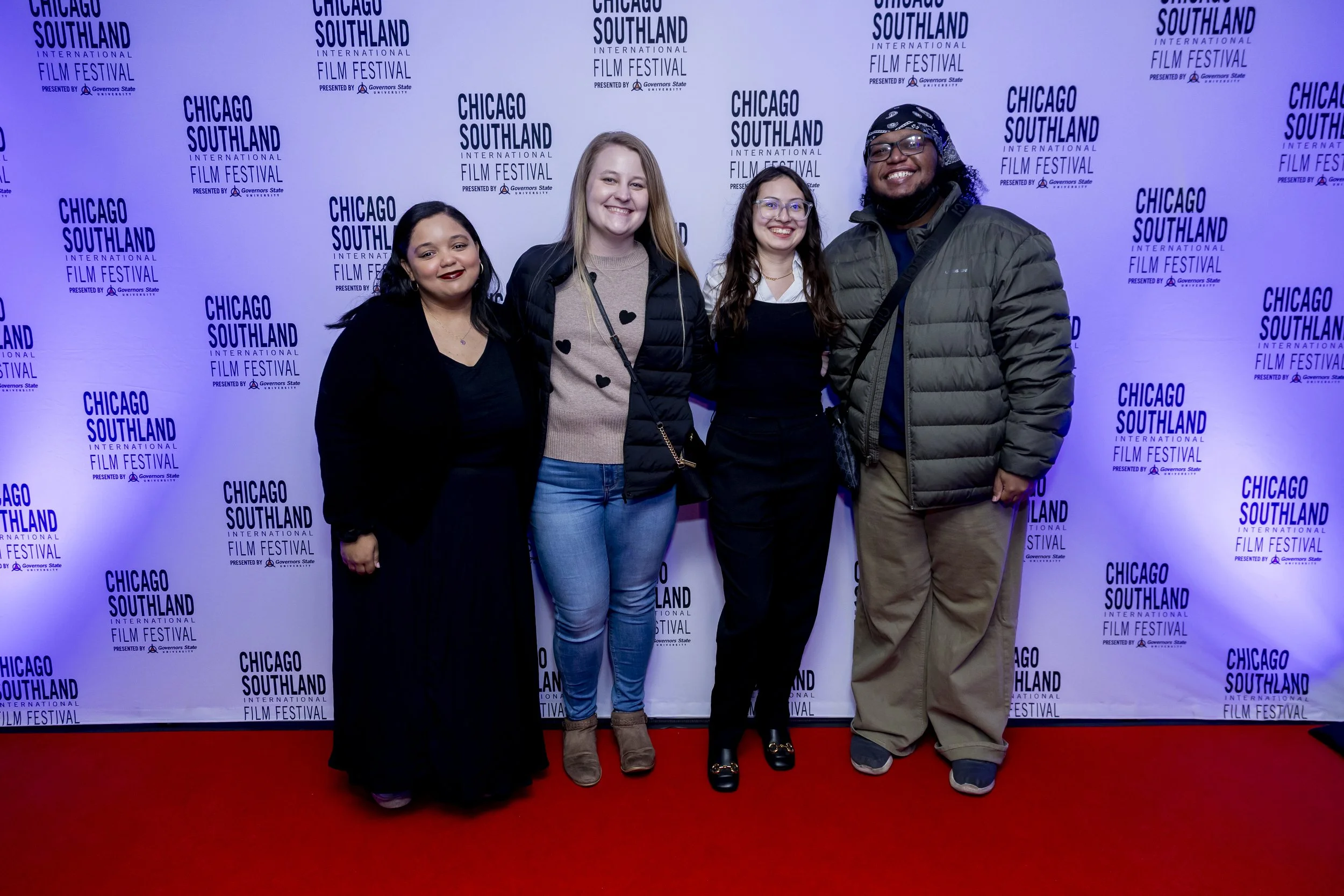 Four people standing in front of a backdrop that reads "Chicago Southland International Film Festival," smiling for a photo. The group includes two women with dark hair, a woman with blonde hair, and a man wearing glasses and a bandana.