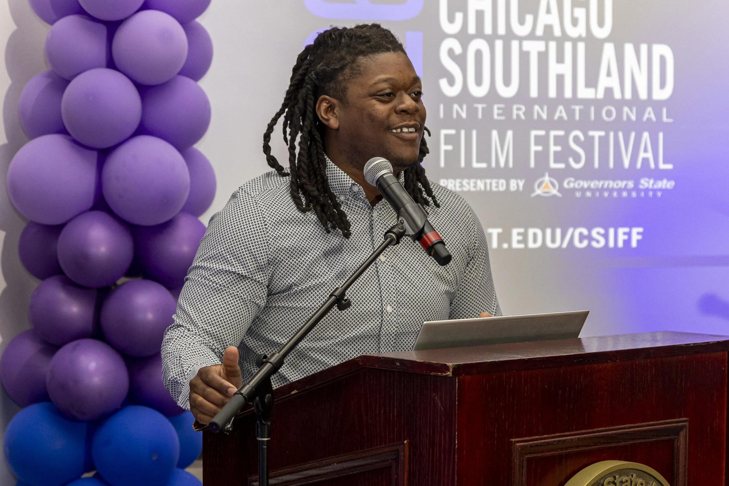 A person with dark skin and dreadlocks is standing at a podium, speaking into a microphone. The background features a wall with purple balloons on the left and a sign that reads 'Chicago Southland International Film Festival' with a logo and addition