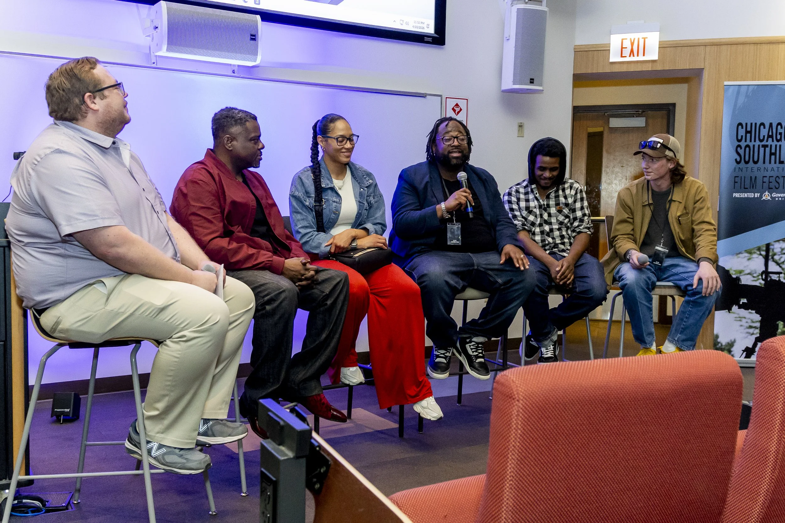 Six diverse people sitting on high chairs on a stage, participating in a panel discussion at the Chicago South International Film Festival, with a screen and exit sign visible in the background.