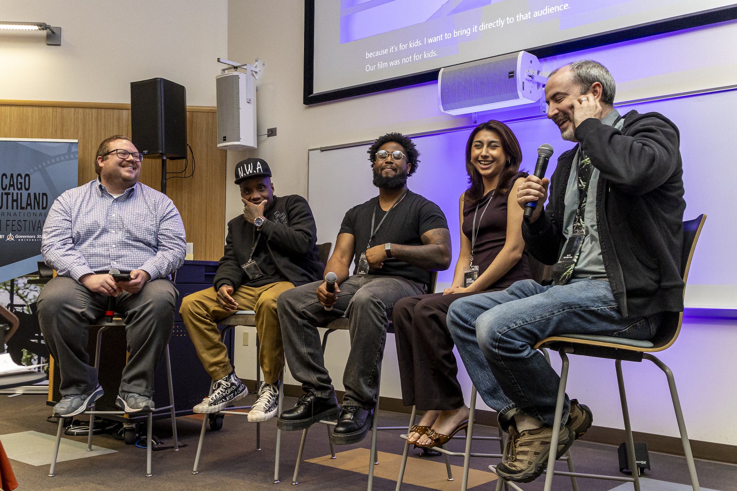 A panel of five diverse people sitting on stage during a discussion at a conference. They are smiling and some are holding microphones. There is a large screen above them with text, speakers mounted on the wall, and a banner with event information.
