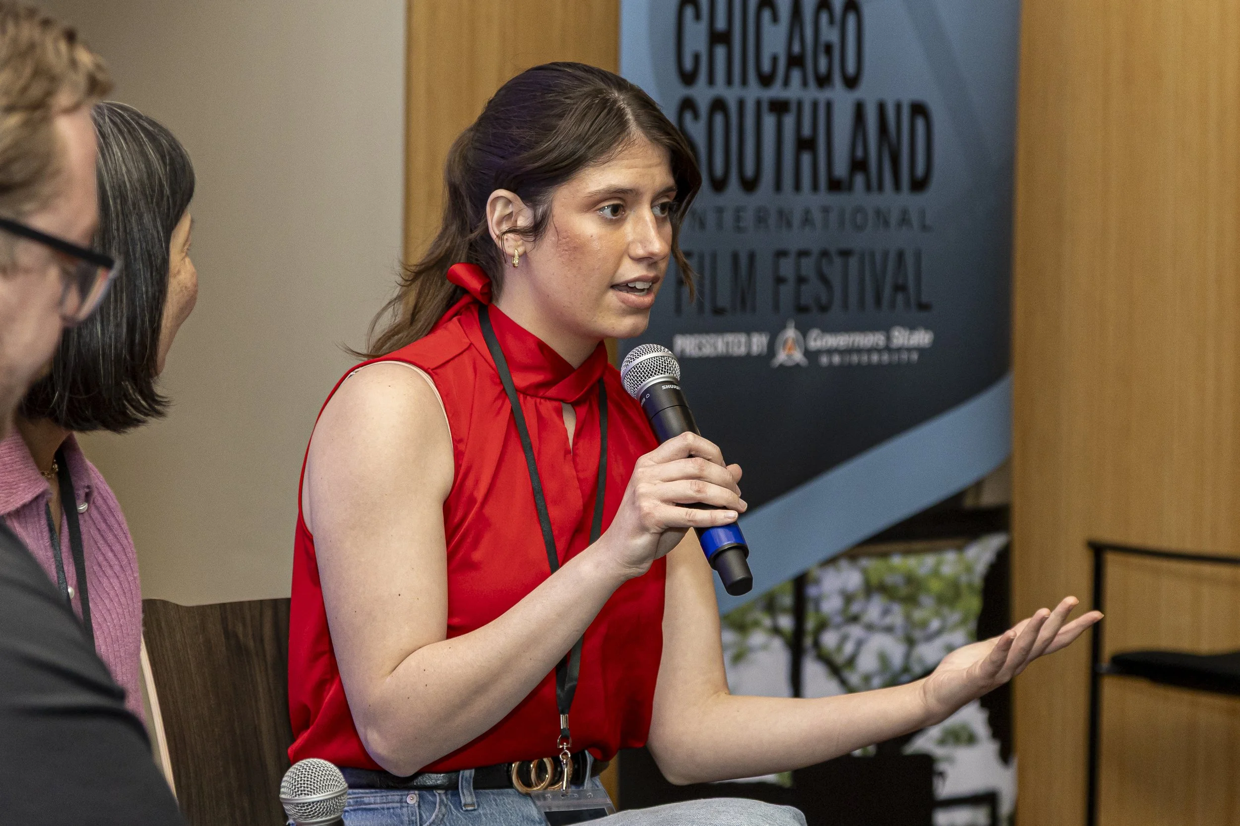 A young woman in a red sleeveless top with a red bow in her hair, speaking into a microphone at a panel or event. A sign in the background reads 'CHICAGO SOUTHLAND INTERNATIONAL FILM FESTIVAL'.