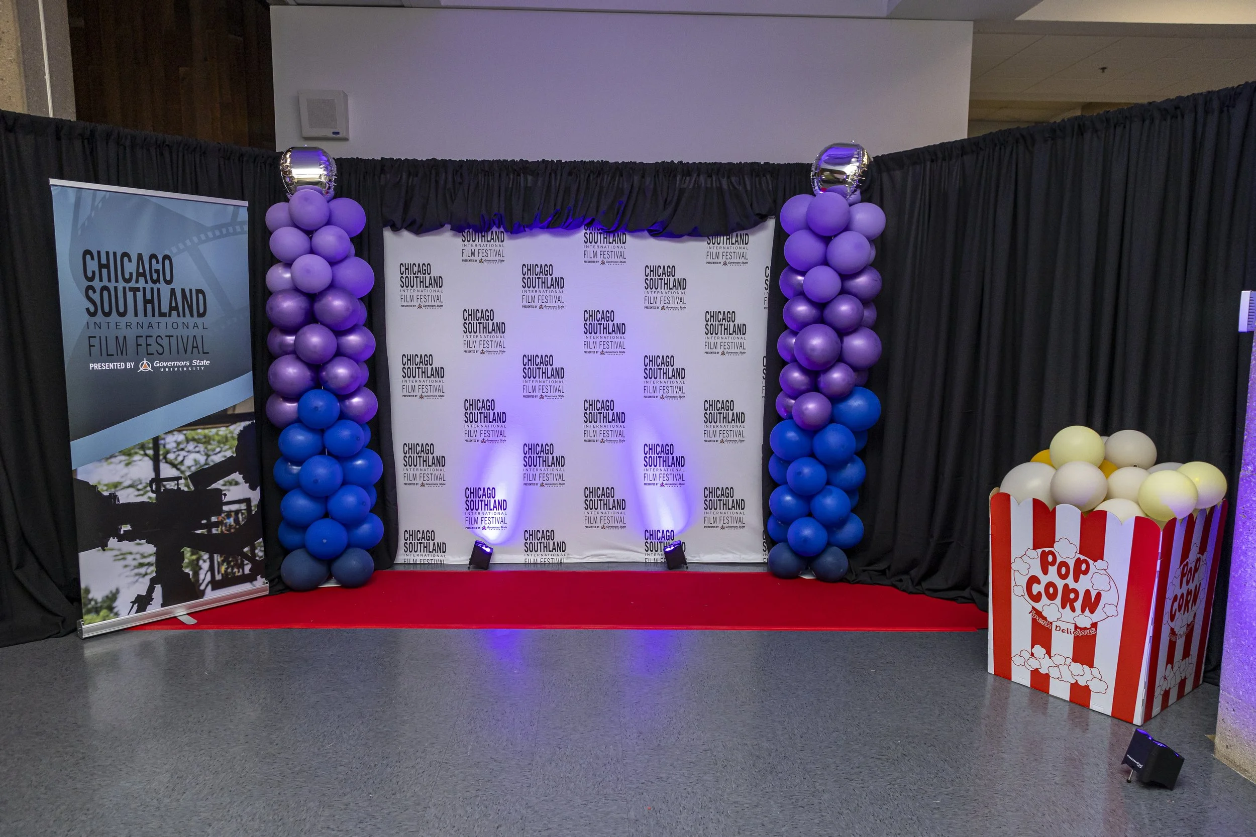 A decorated stage area for the Chicago Southland International Film Festival with black curtains, purple and blue balloon columns on either side of a backdrop with the festival logo, and a popcorn stand on the right side.