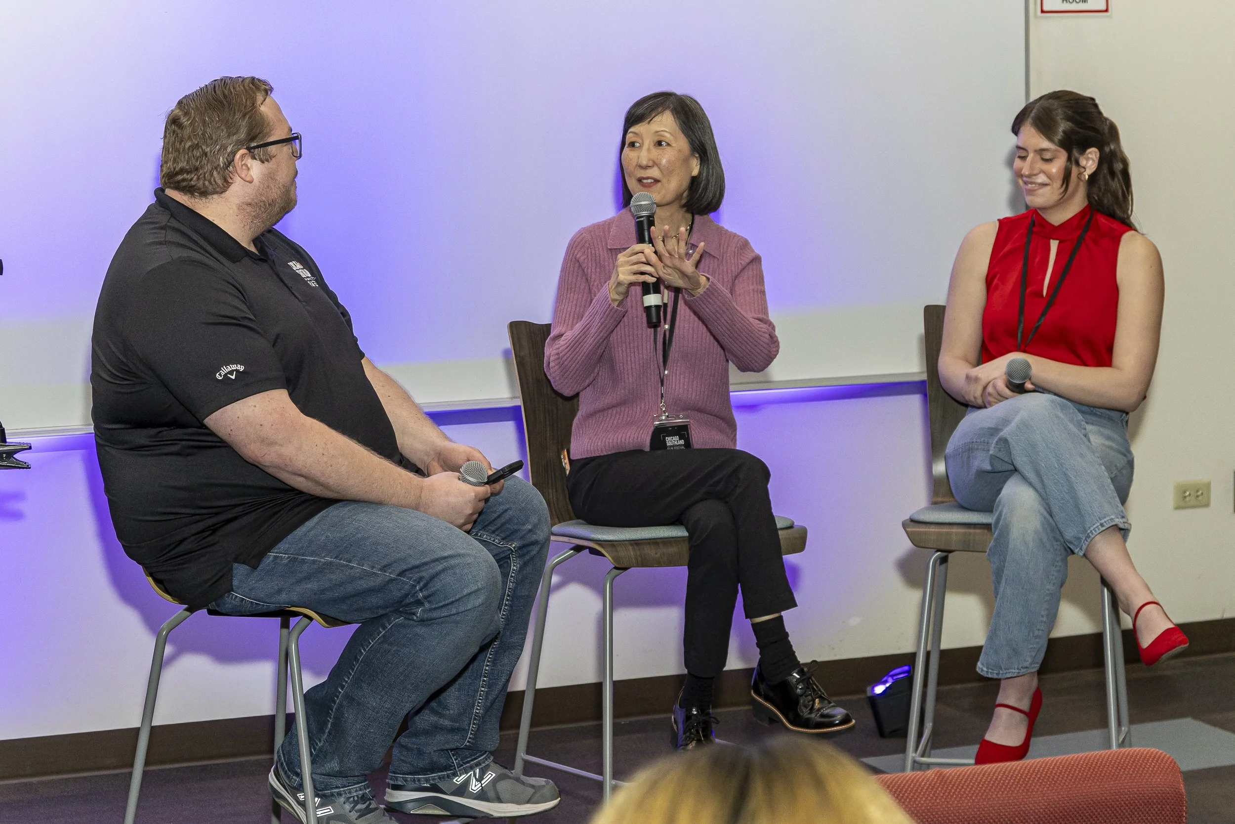 Three people are engaged in a panel discussion or interview. The person in the middle is speaking into a microphone, while the person on the left is listening and holding a microphone. The person on the right is smiling, sitting cross-legged, and hol