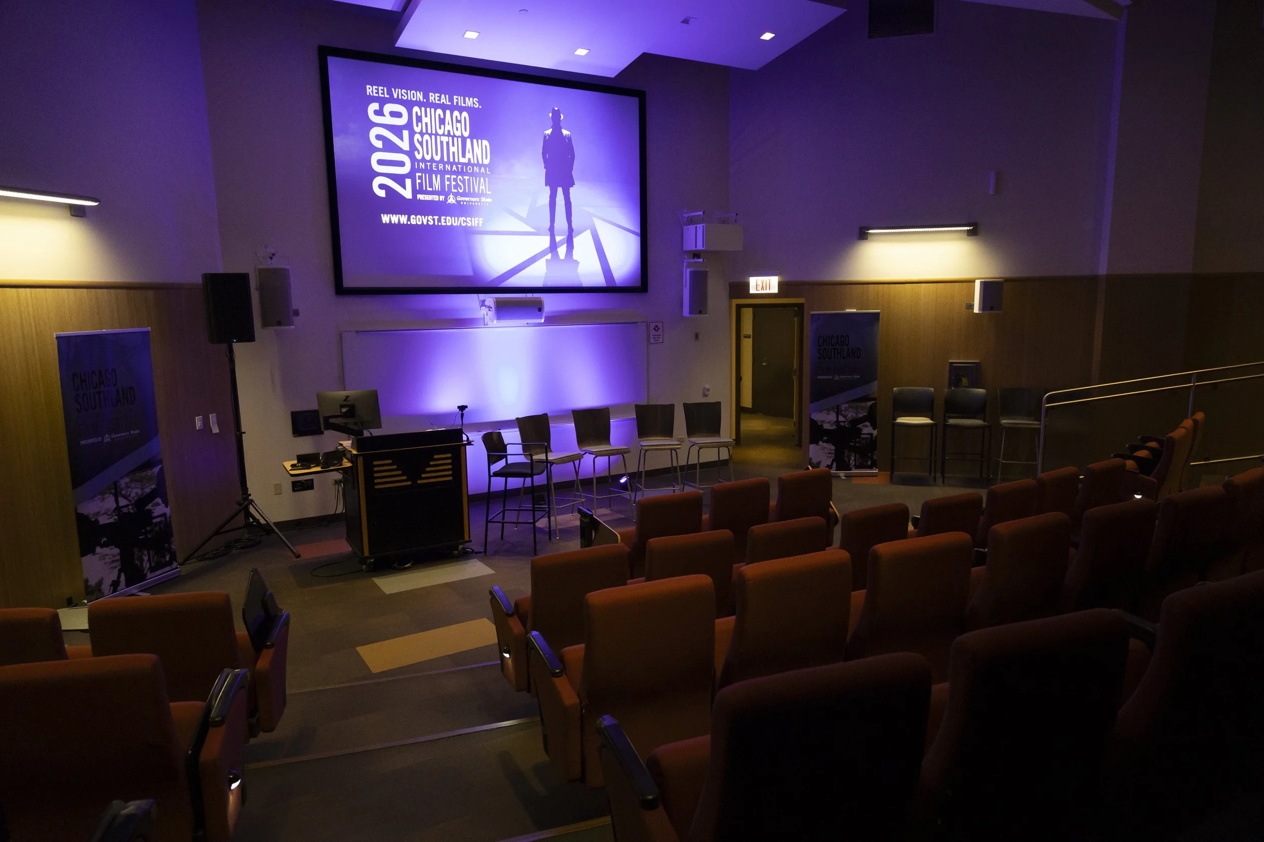 A theater or conference room with a large screen displaying information about the 2026 Chicago Southland International Film Festival, with rows of red seats and purple ambient lighting.