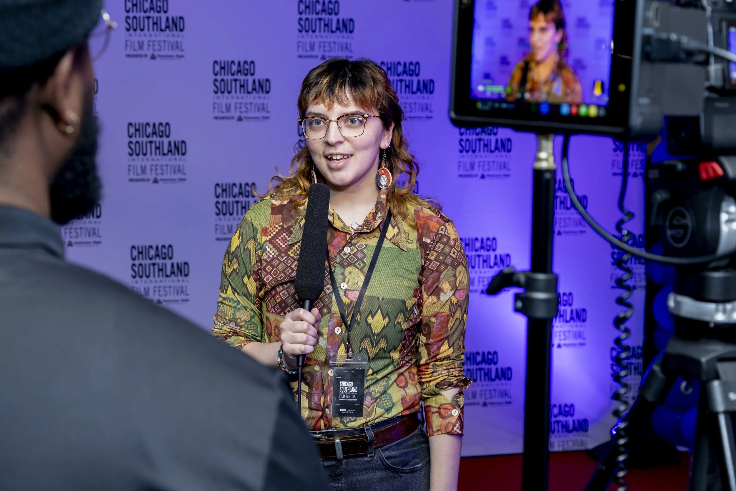 A woman with glasses and a patterned shirt being interviewed with a microphone at the Chicago Southland Film Festival, with a backdrop displaying the festival's logo.