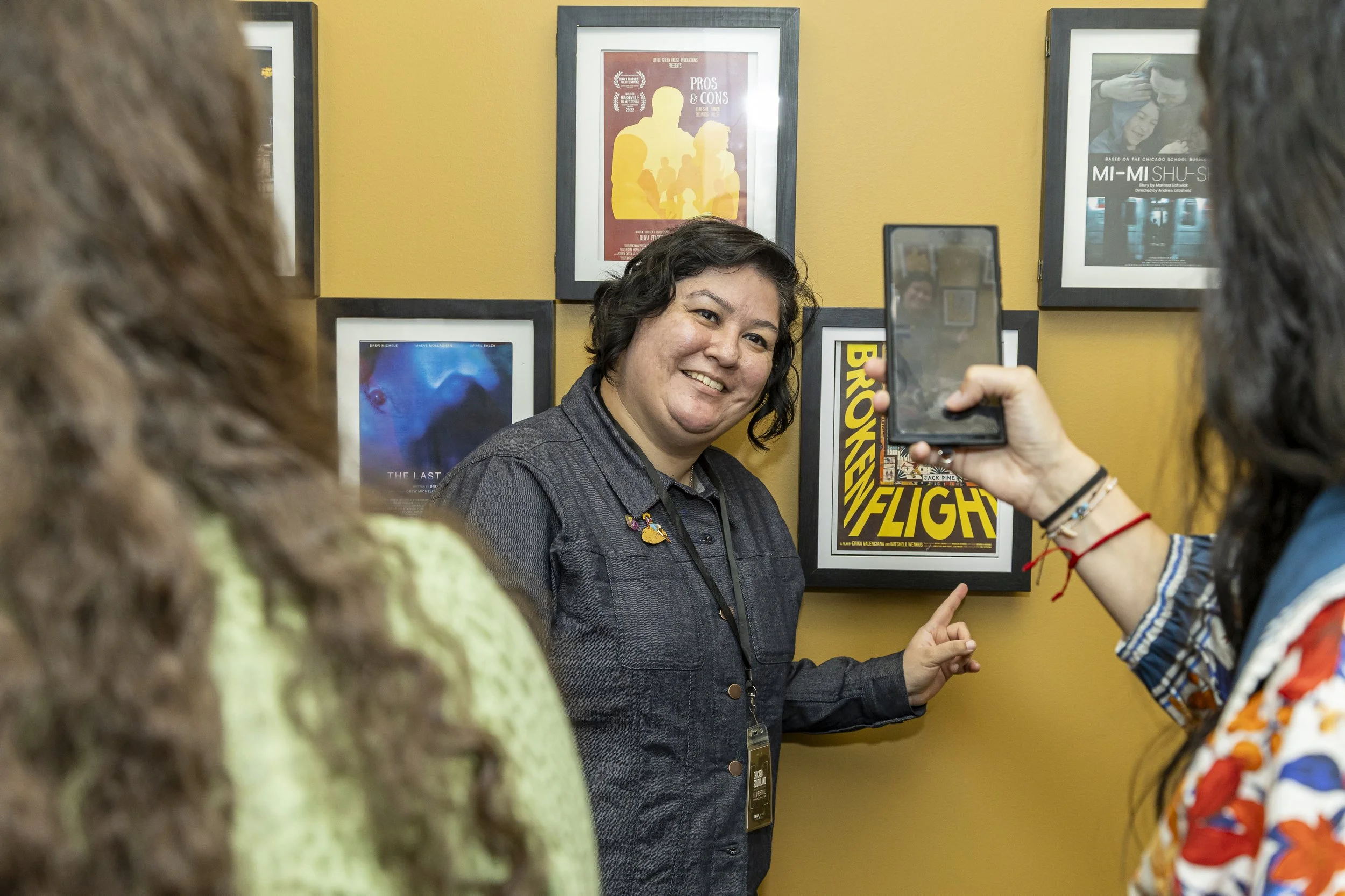 A woman with curly hair and a dark shirt is smiling while being interviewed or photographed by others in front of a yellow wall with framed posters.