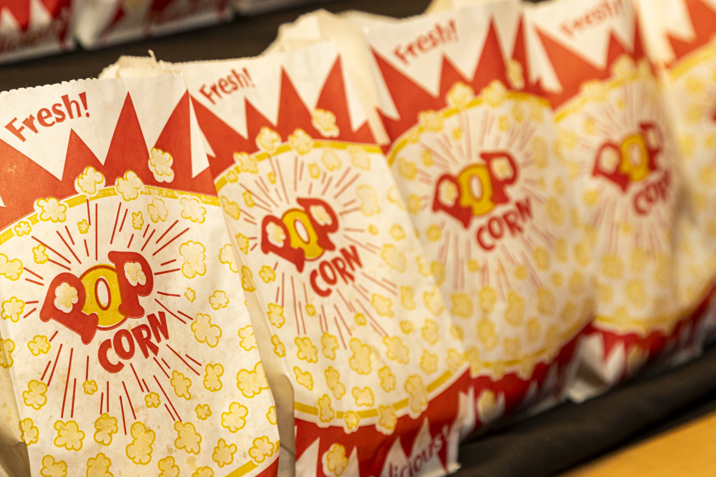 Close-up of popcorn bags with red and yellow design and the words 'Fresh!' and 'Popcorn' printed on them, arranged on a shelf.