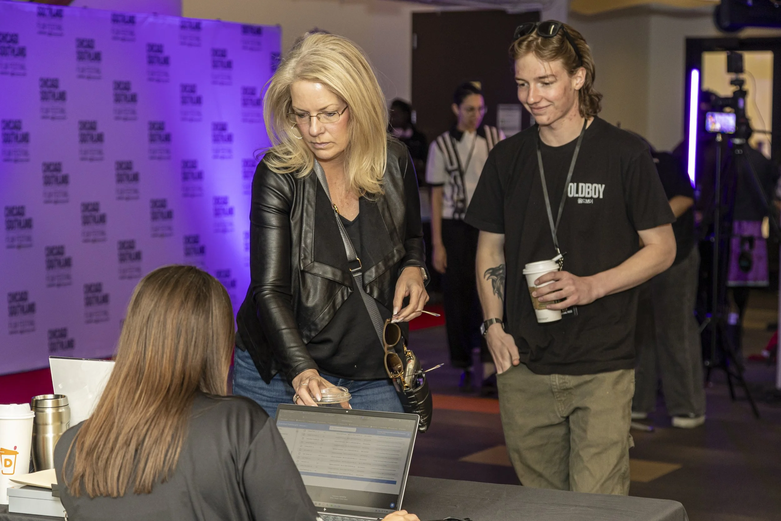 Two women, one with blonde hair and glasses and the other with brown hair and sunglasses on her head, are standing at a table in a conference or event setting. The blonde woman is looking at a piece of paper on the table, while the brunette woman is 