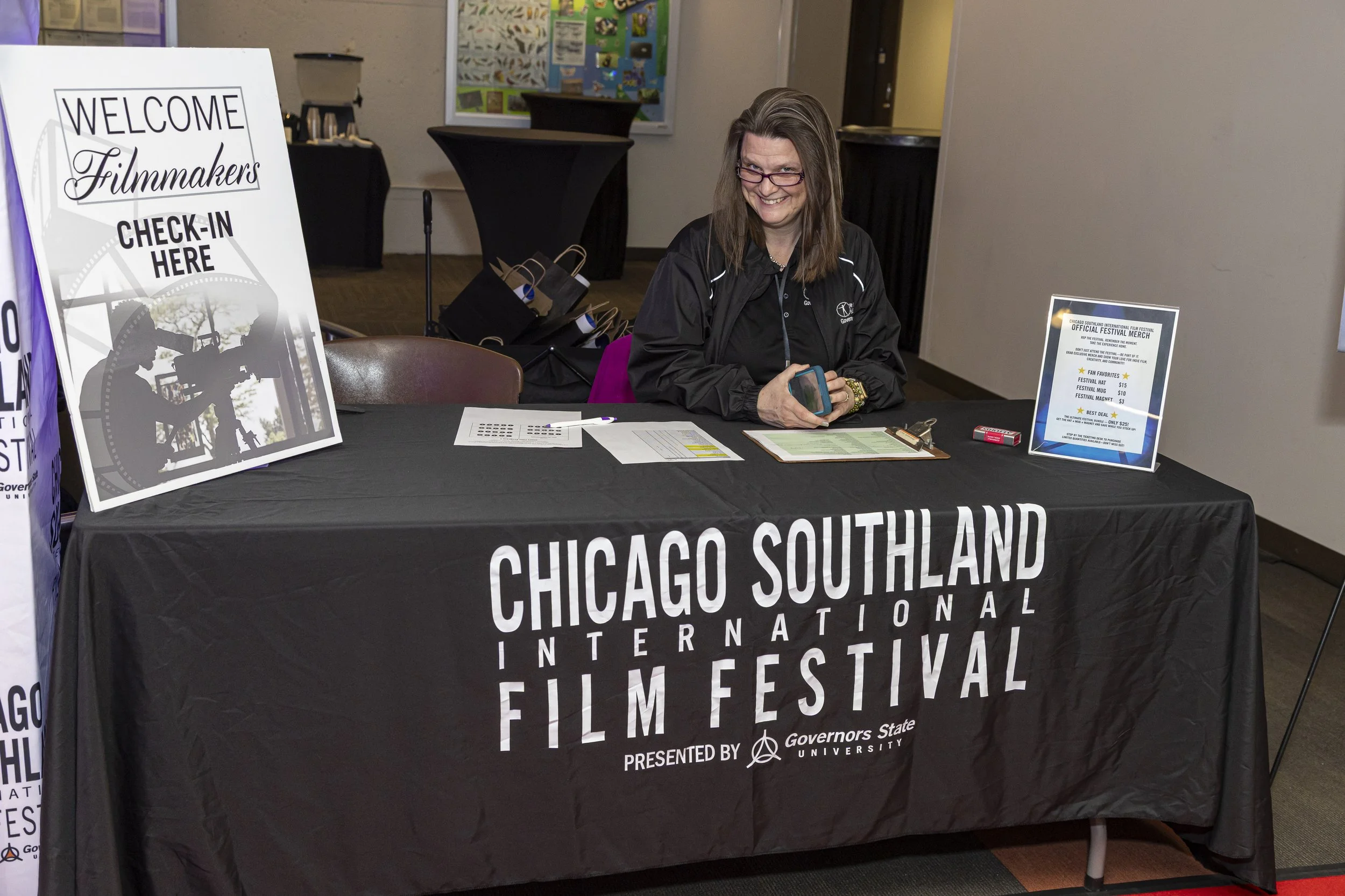 A woman sitting behind a table with a black tablecloth that reads: 'Chicago Southland International Film Festival.' She is smiling, holding a phone, wearing glasses and a black jacket. On the table are papers, a pen, and a small sign about festival m