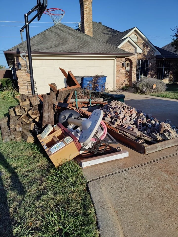 A residential driveway with a large pile of construction debris including wood logs, bricks, and household items like a baby chair and treadmill. In the background, a garage and a house with a basketball hoop on the driveway are visible.