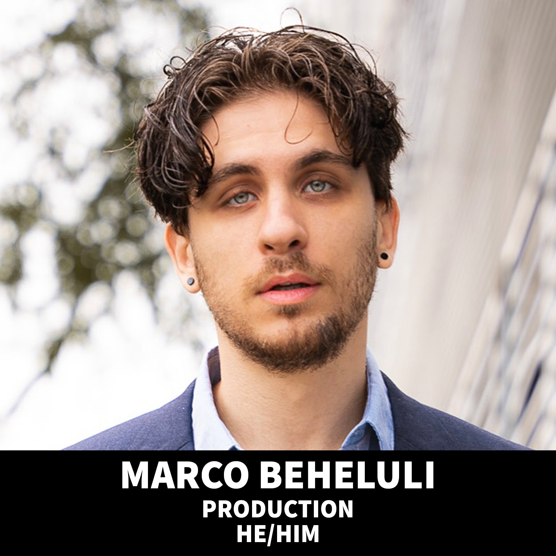 A young man with a beard, blue eyes, and earrings, wearing a black cap backwards and a black t-shirt, standing against a black background.