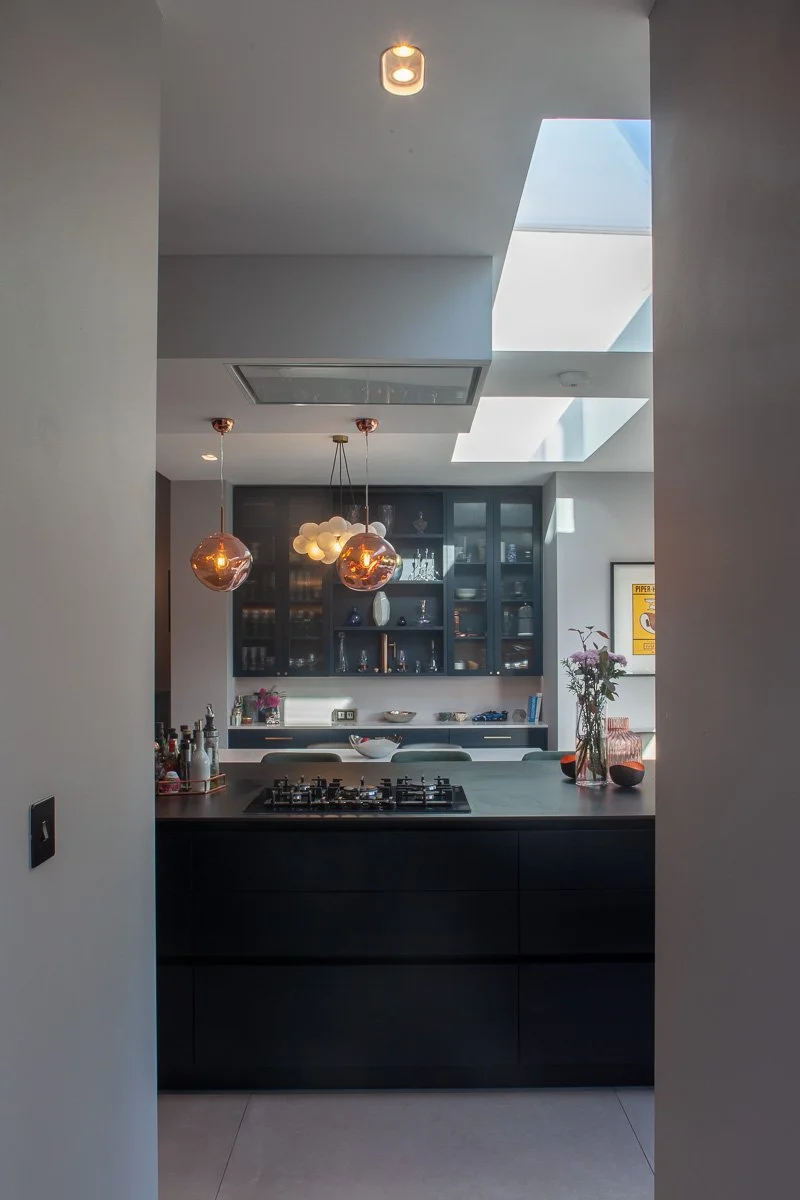 Dark fitted kitchen viewed through a wide opening with island hob, display cabinetry and pendant lighting