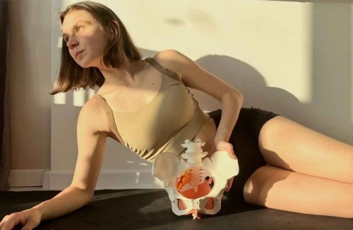 Woman in a tank top and shorts holding a pelvic bone model while reclining on a yoga mat in a sunlit room.