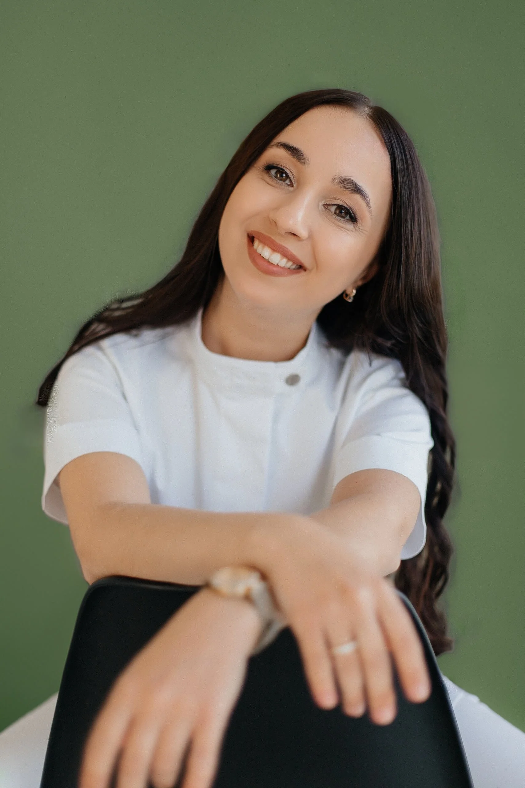 A smiling woman with long brown hair wearing a white shirt, leaning on a chair with a green background.