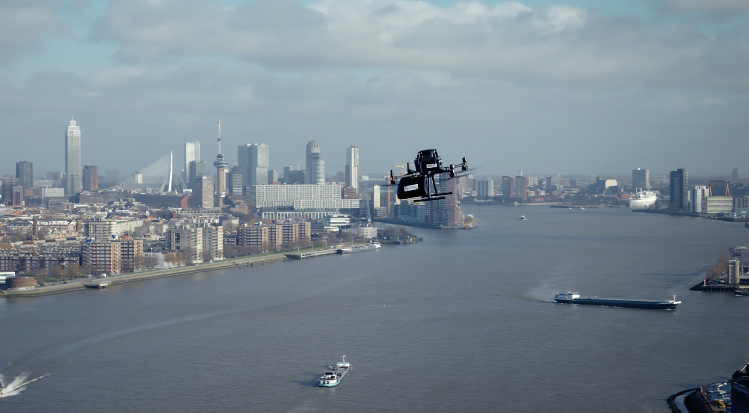 Een drone vliegt over de rivier met de skyline van een grote stad op de achtergrond, met hoge gebouwen, boten op de rivier en een gekleurde lucht met wolken.