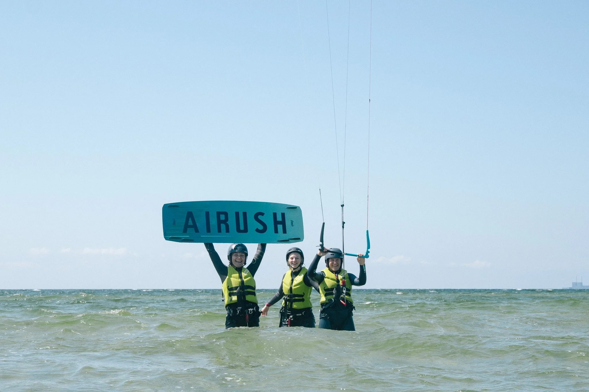 Två personer är på stranden, en av dem håller i ett annat person som är med på en kitesurfingaktivitet. Kitesurfingutrustning och vågor syns i havet.