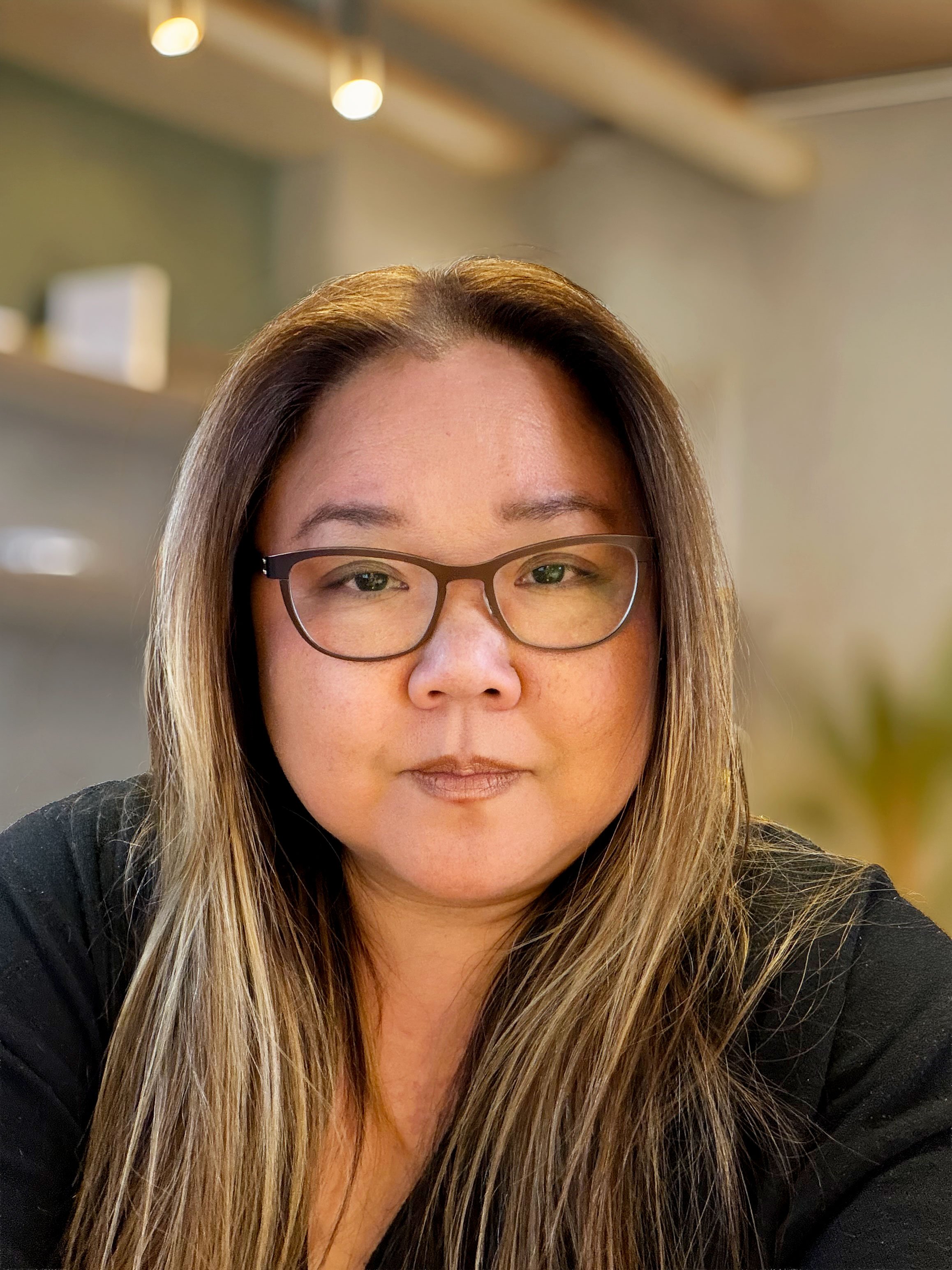 A woman with long brown hair, wearing glasses and a green top, standing outdoors with cityscape and blue sky in the background.