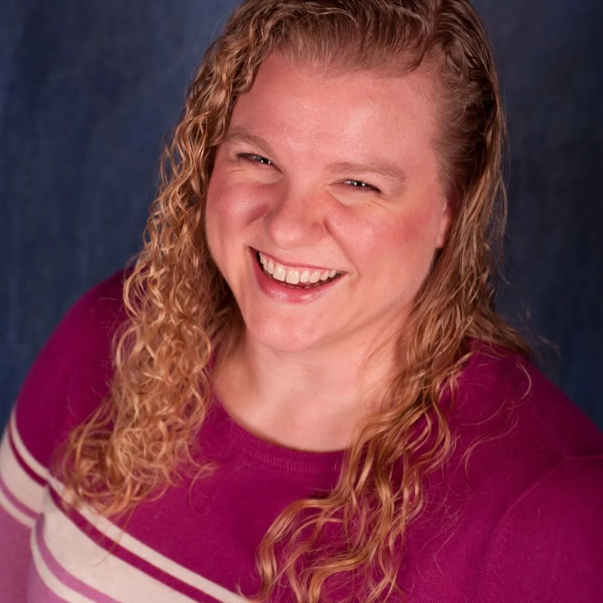 A woman with long, curly red hair wearing a maroon shirt smiles with a joyful expression against a dark background.