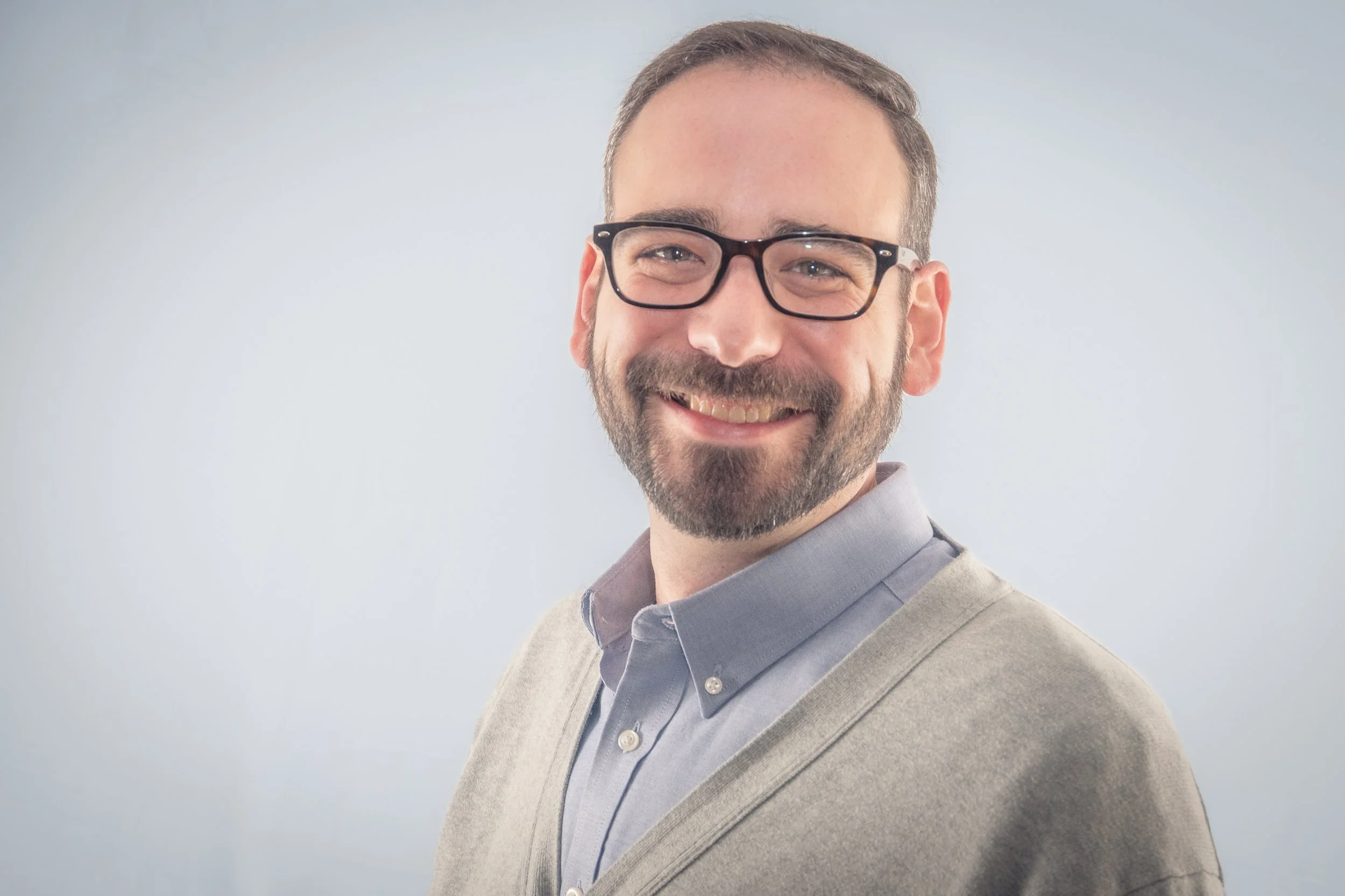 Portrait of a smiling man with glasses, wearing a light grey blazer and a blue shirt, against a plain light background.