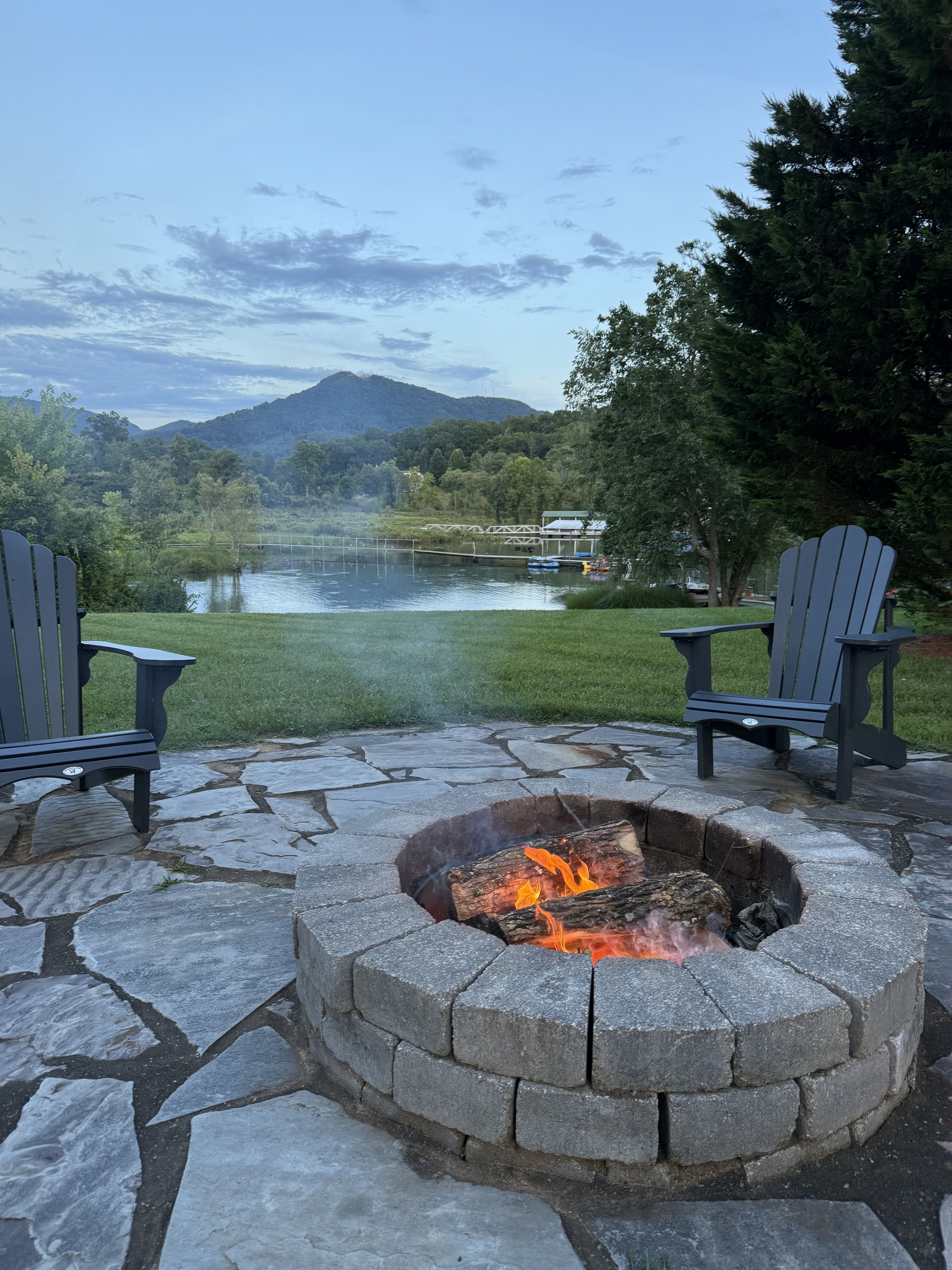 View of a backyard with a fire pit and two chairs, overlooking a river, trees, and mountains in the distance during early evening or dawn.