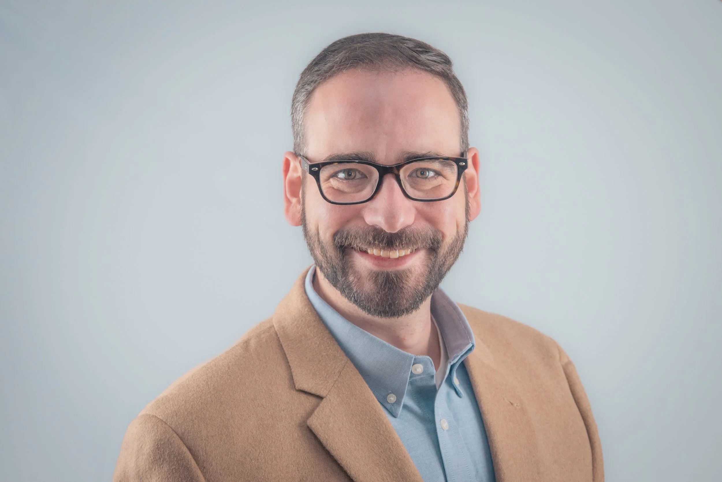 Portrait of a smiling man with glasses, a beard, wearing a light blue shirt and a brown blazer, against a plain background.