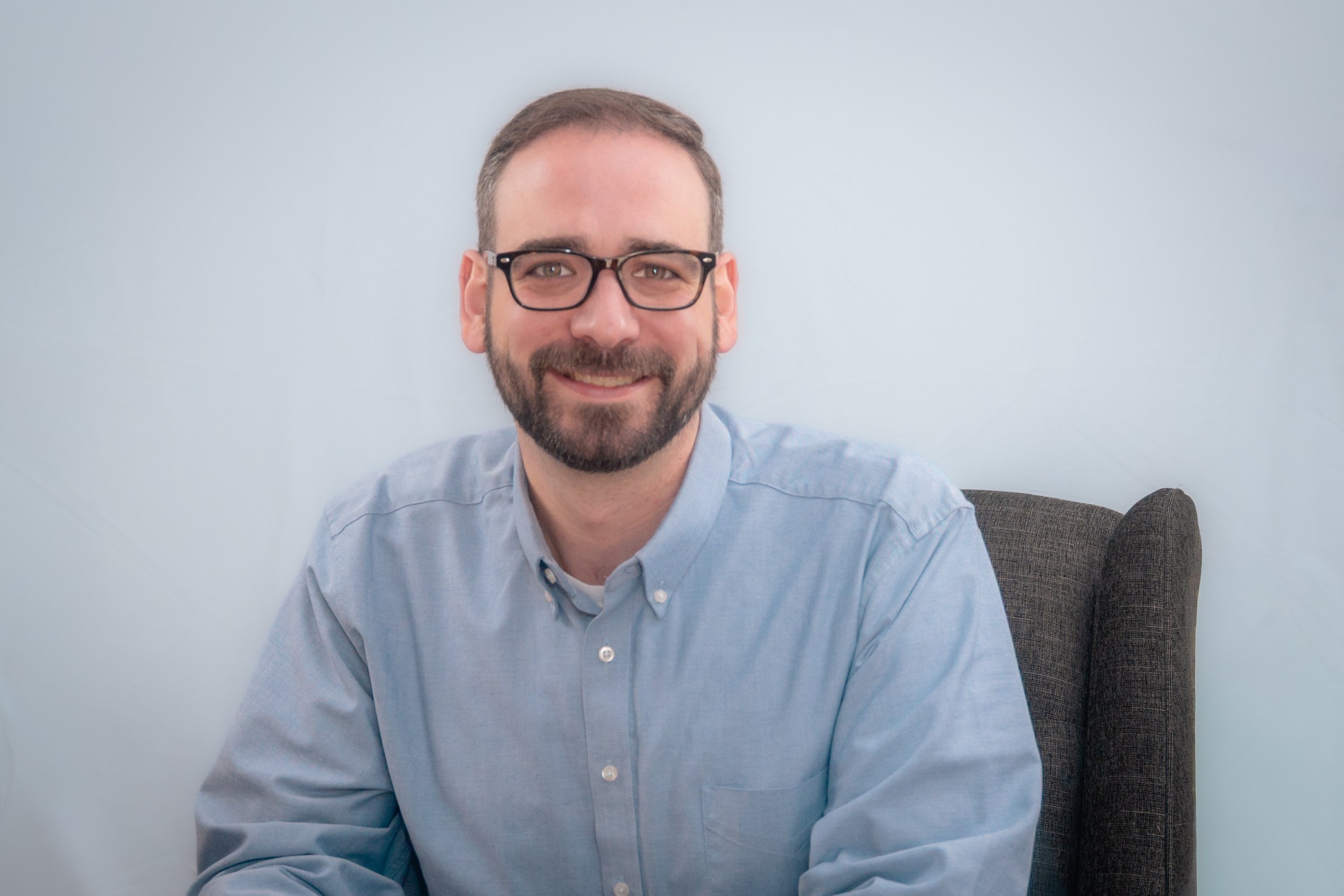 A man with glasses and a beard smiling, sitting in a gray chair against a plain light blue wall, wearing a light blue collared shirt.