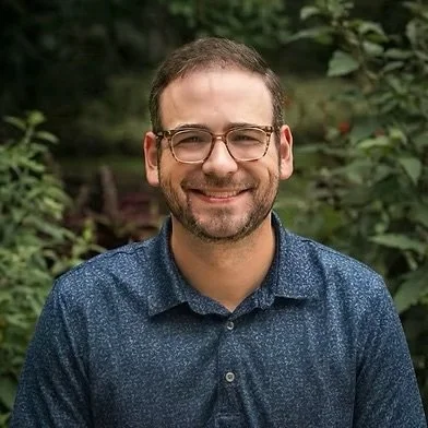 A man with glasses and a beard smiling outdoors, wearing a blue collared shirt, surrounded by greenery.