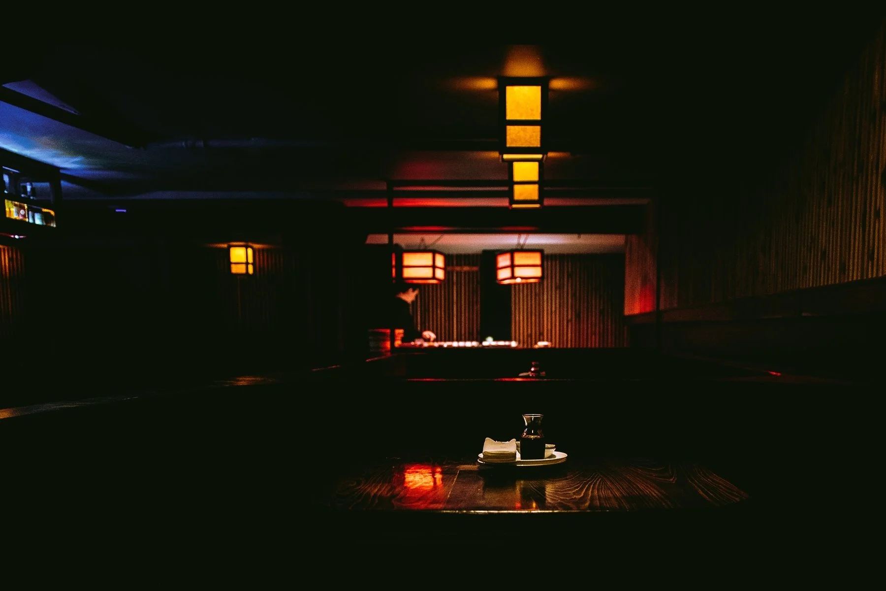 A dimly lit restaurant with dark wood furnishings, hanging soft yellow and red lantern-style lights, a table set with a small plate, napkin, and soy sauce bottle, and a person in the background near a buffet or serving area.