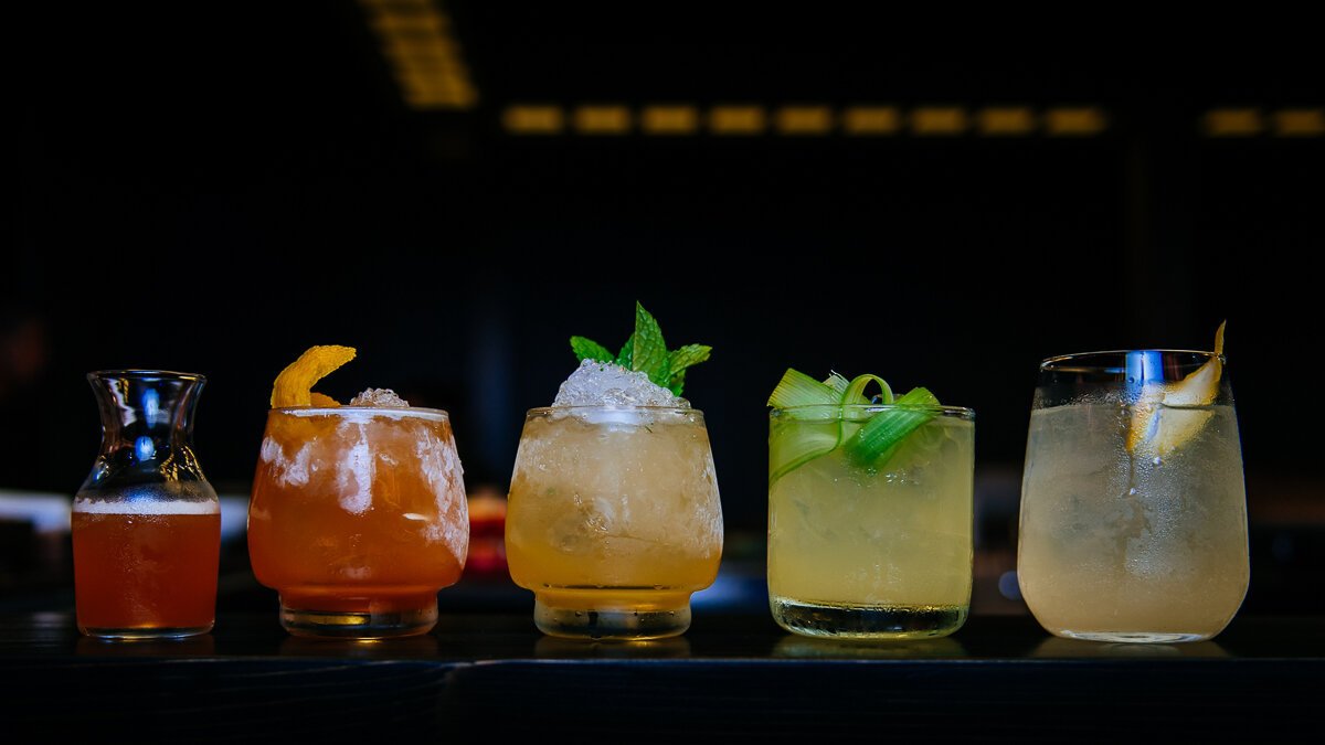 Five cocktail glasses with various drinks and garnishes on a dark bar surface.