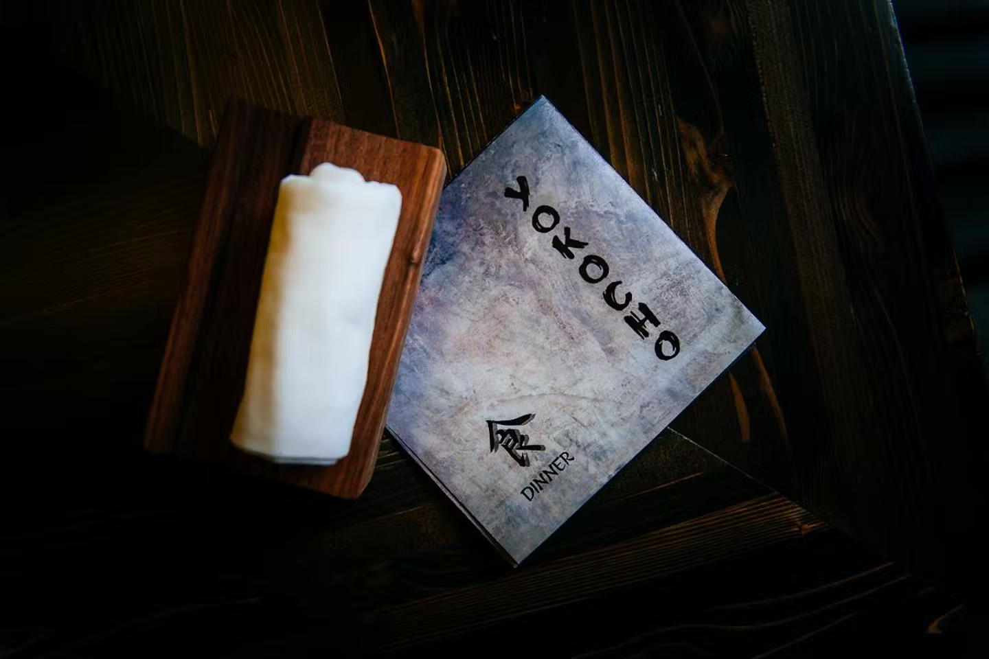 A menu card titled 'DINNER' with Japanese characters and a candle on a wooden holder, placed on a dark wooden table.