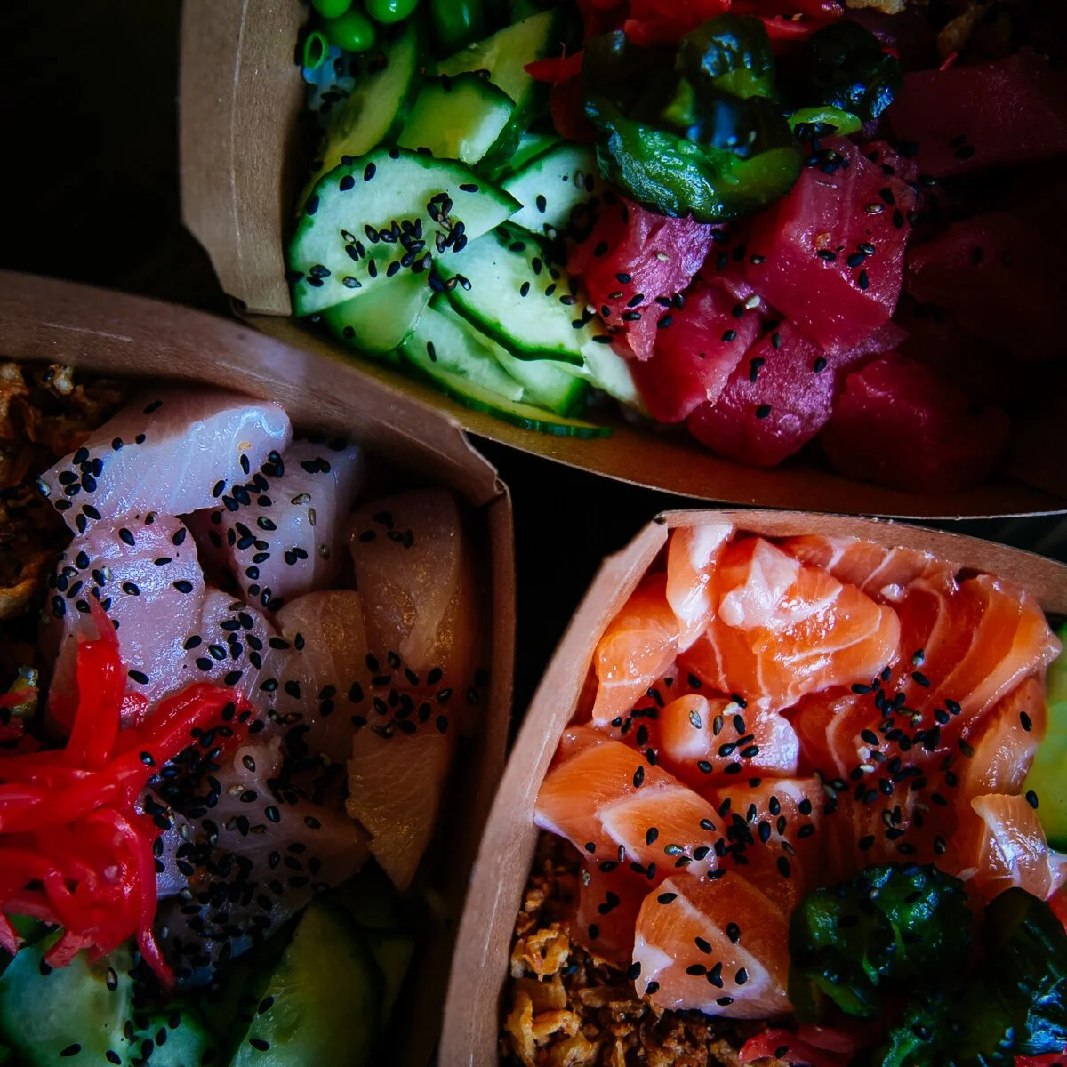 Assorted poke bowls with sliced cucumber, avocado, fried shallots, edamame and diced raw fish, topped with black sesame seeds.