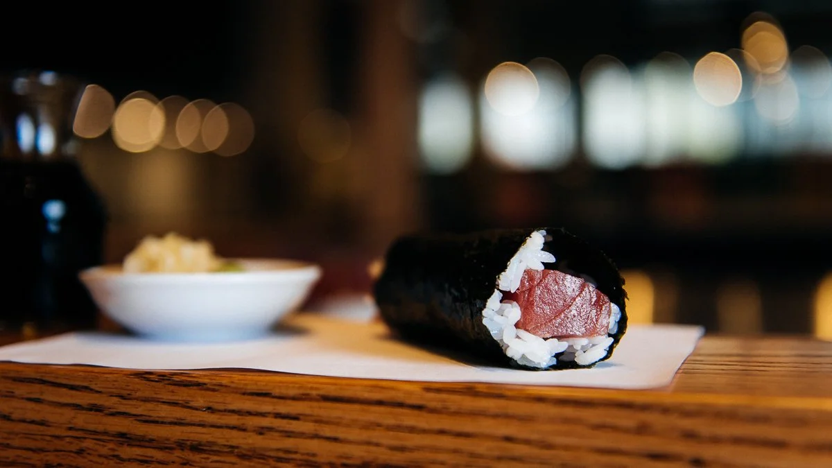 Close-up of a sushi hand roll with tuna and rice, wrapped in seaweed on a wooden surface with a bowl of wasabi in the background, out of focus.