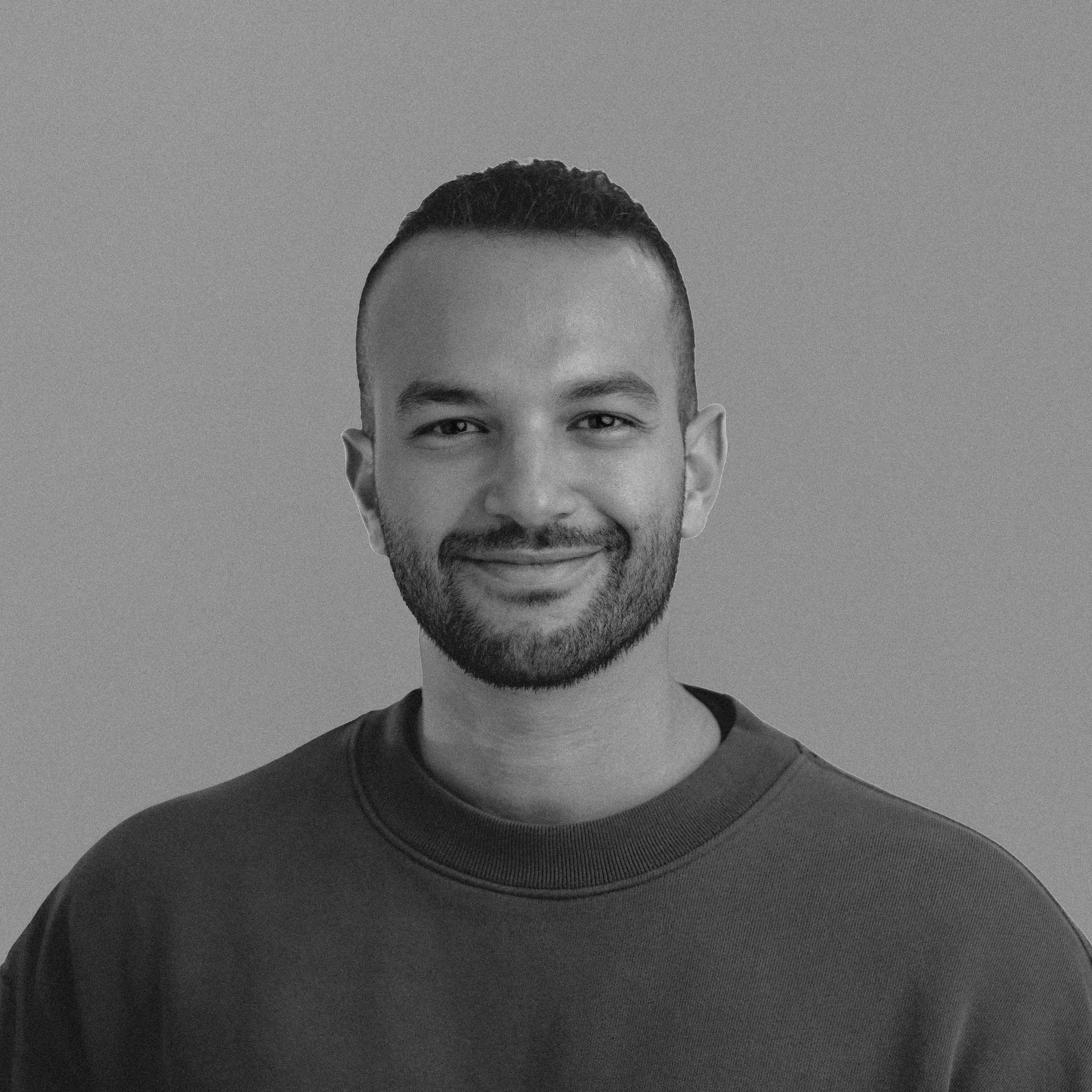 Close-up of a young man smiling, with short dark hair, a beard, and wearing a dark T-shirt, against a plain light background.