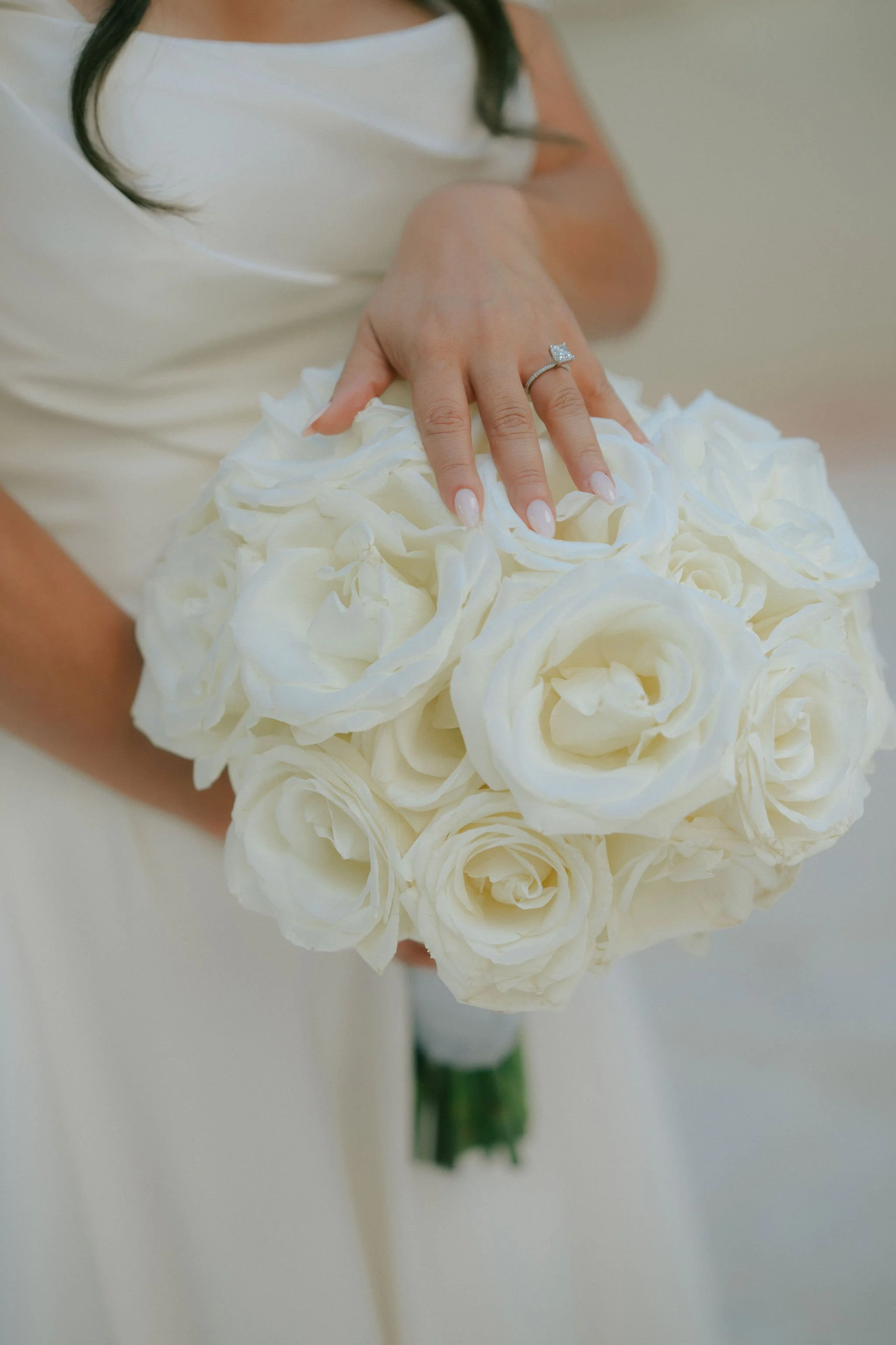 A woman in a white dress holds a bouquet of white roses, with her left hand resting on top, showing a diamond ring on her ring finger.