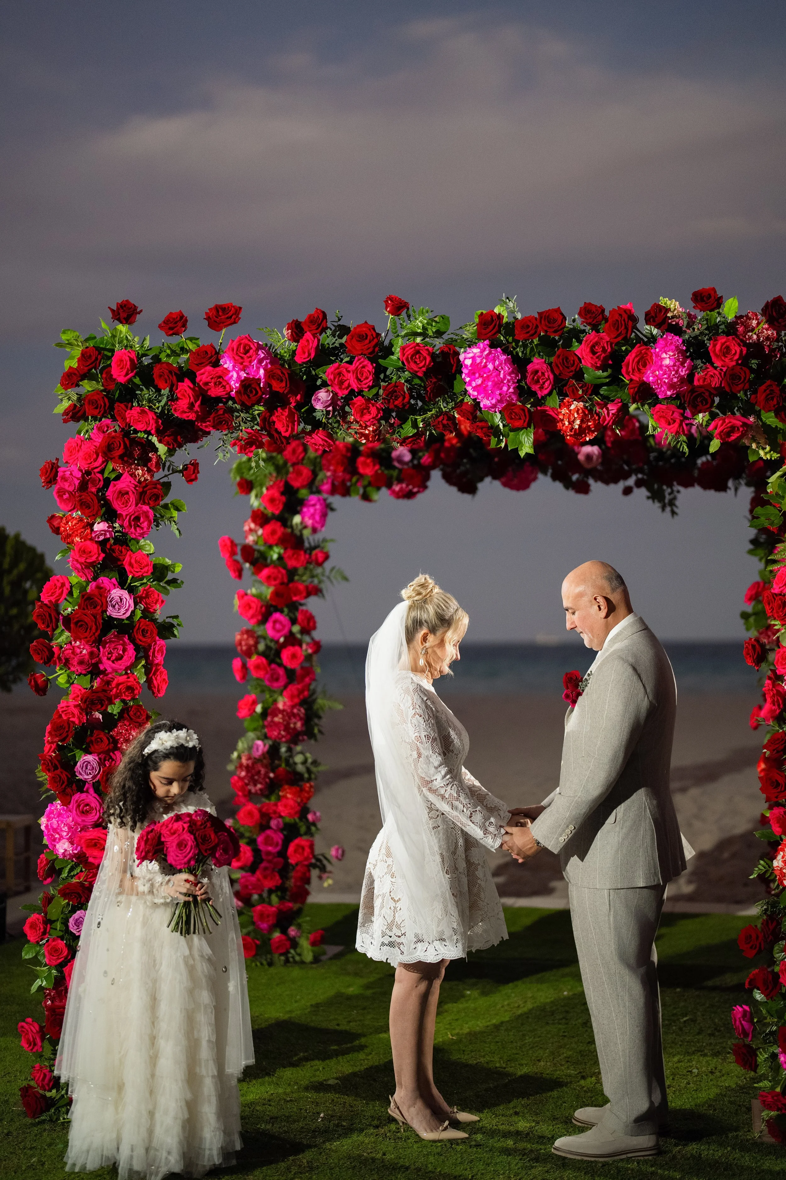 A couple getting married outdoors under a floral arch, with a young girl holding a bouquet of flowers nearby.