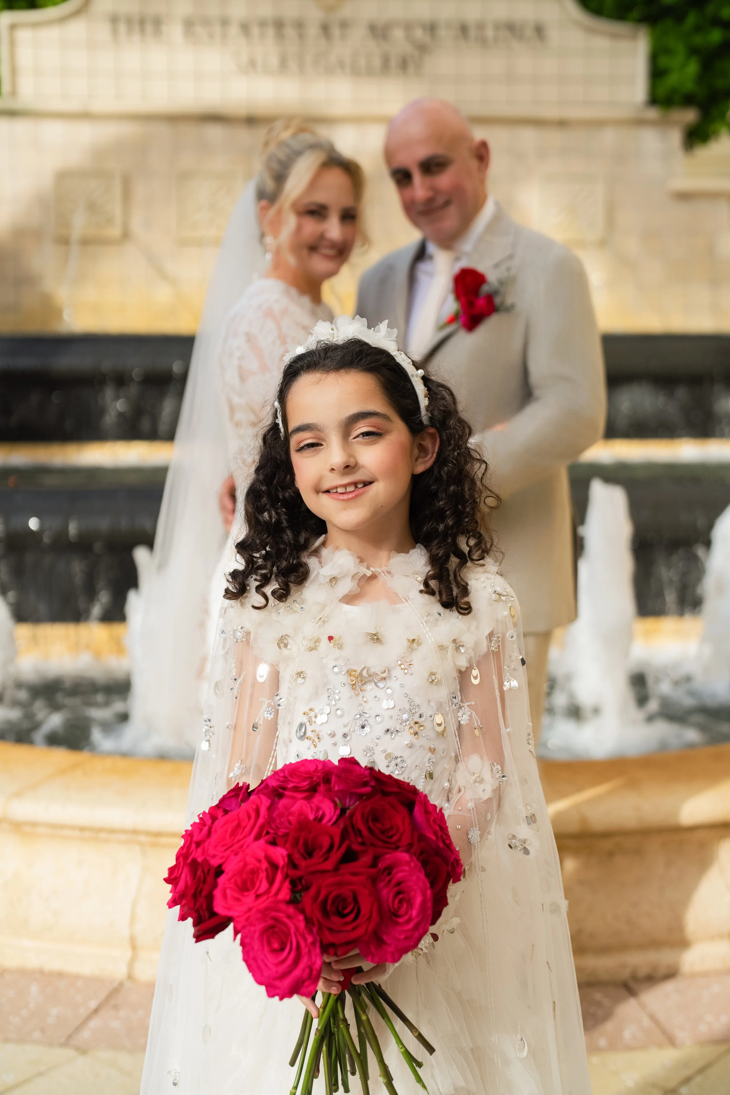 A young girl in a white dress with sequins holds a bouquet of red roses and stands in front of a fountain. Behind her, a bride in a lace wedding gown with a veil and a groom in a beige suit with a red boutonniere are smiling, blurred in the backgroun