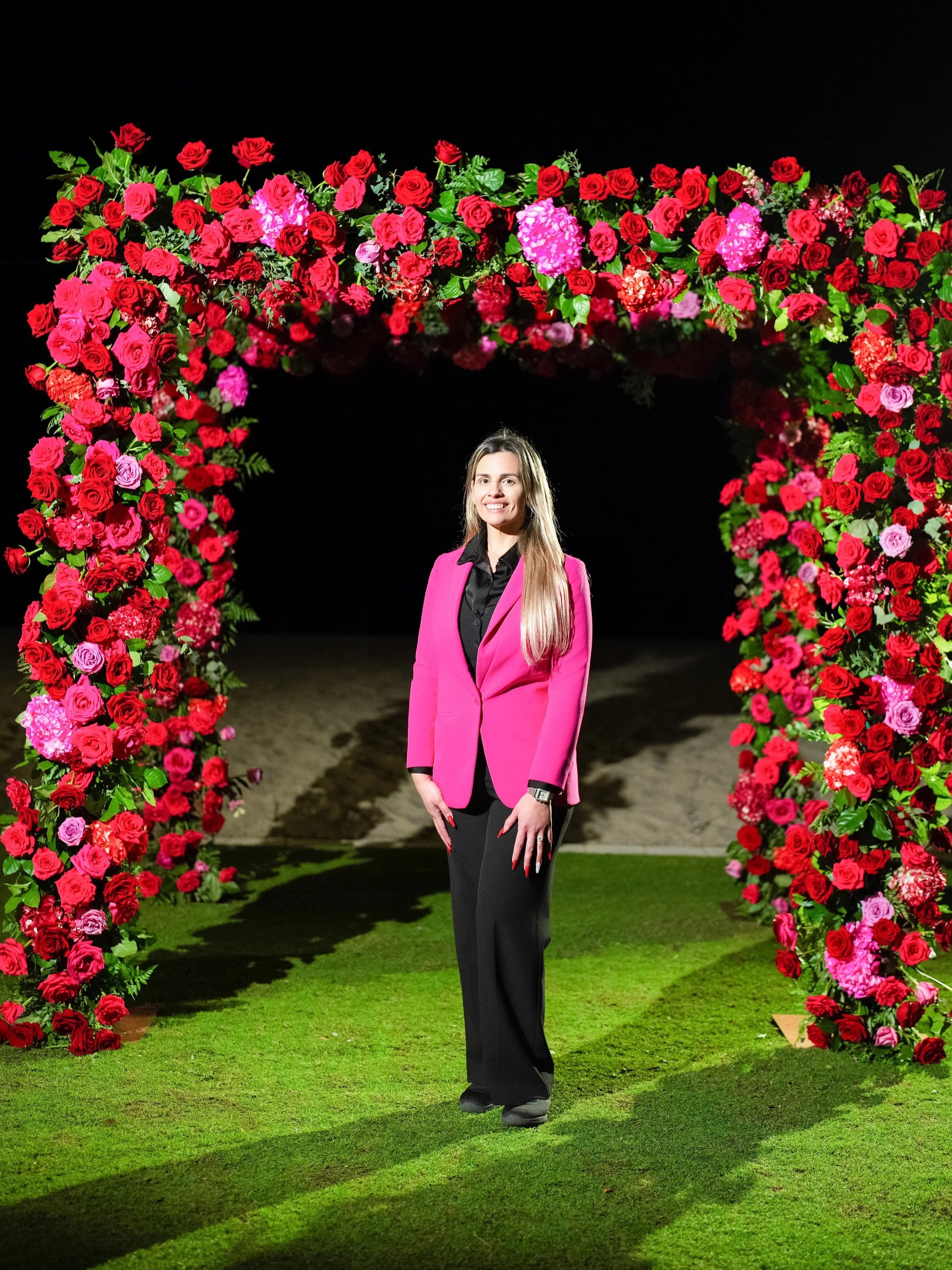 Woman in a pink blazer and black pants standing under a floral arch made of red and pink roses on a green grass field at night.