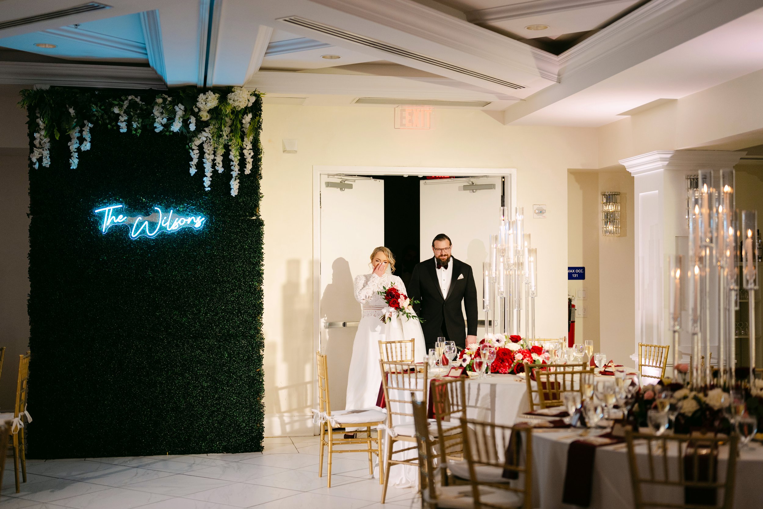 Bride and groom walking into the reception area, decorated with floral arrangements and a neon sign that reads 'The Wilkins'.