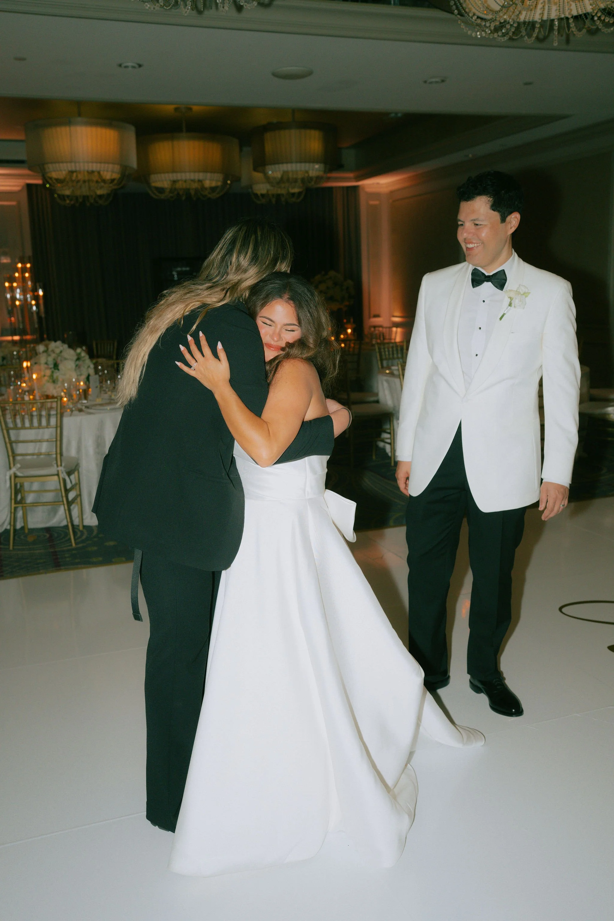 Two women hugging, one in a white dress, and a man in a tuxedo, at a wedding reception.