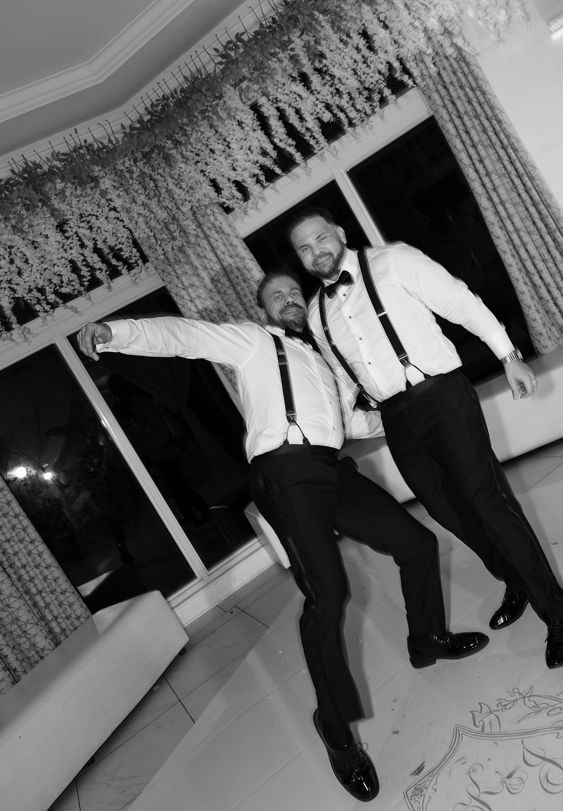 Two men dressed in tuxedos and suspenders dancing and having fun at a decorated indoor event.