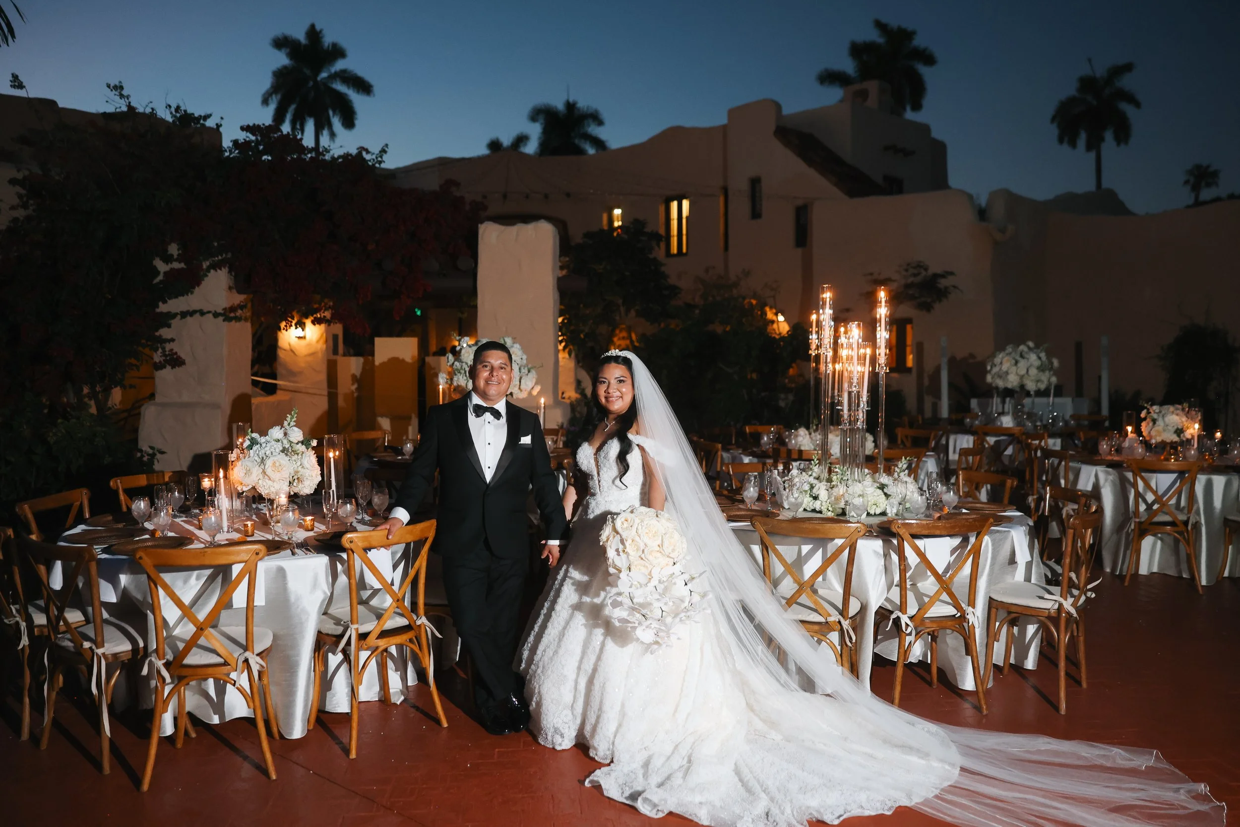 A newlywed couple in wedding attire standing at a decorated reception tables outdoors during nighttime, with tables adorned with white floral arrangements and candles.