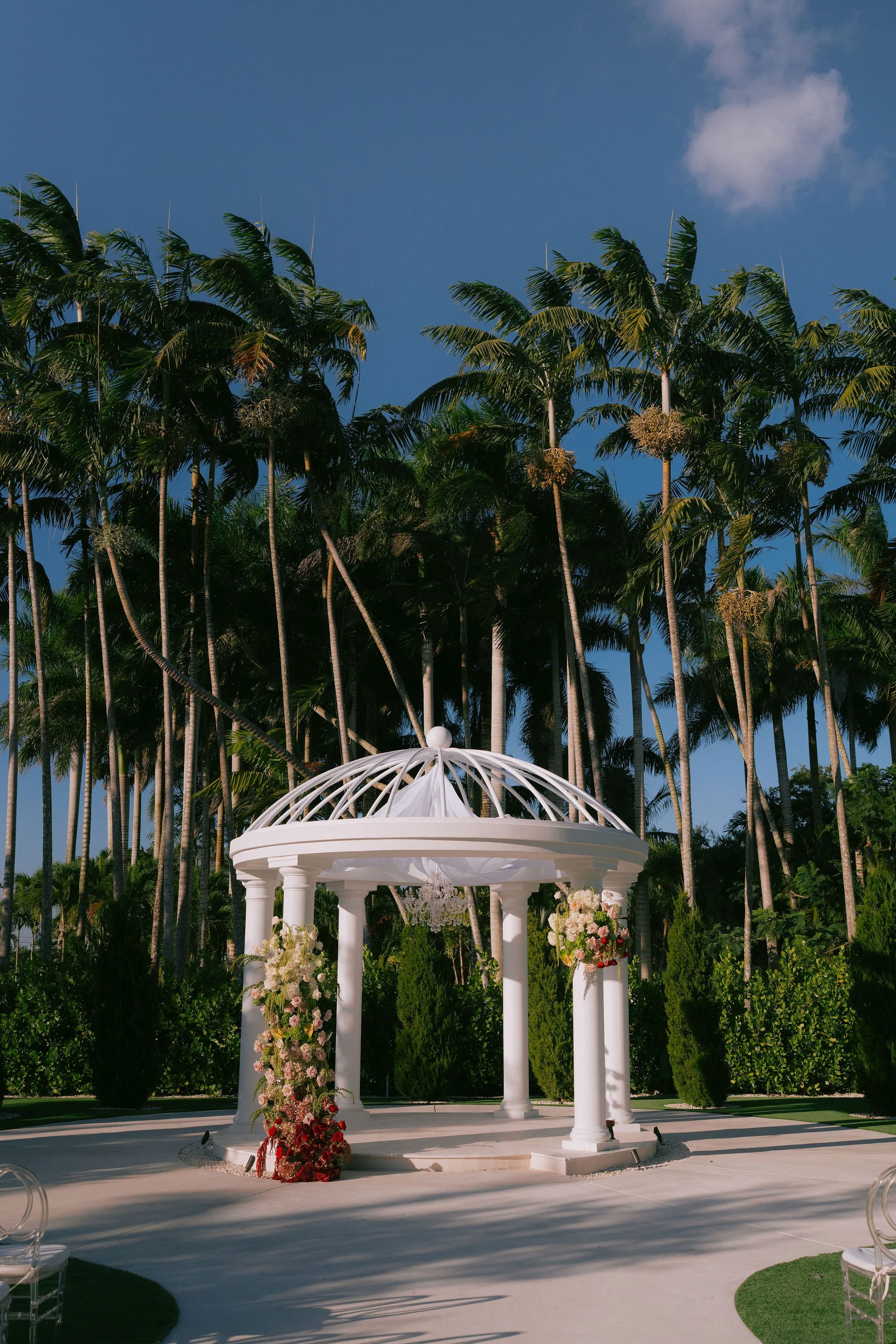 Decorative wedding gazebo with floral arrangements in front of tall palm trees on a sunny day.