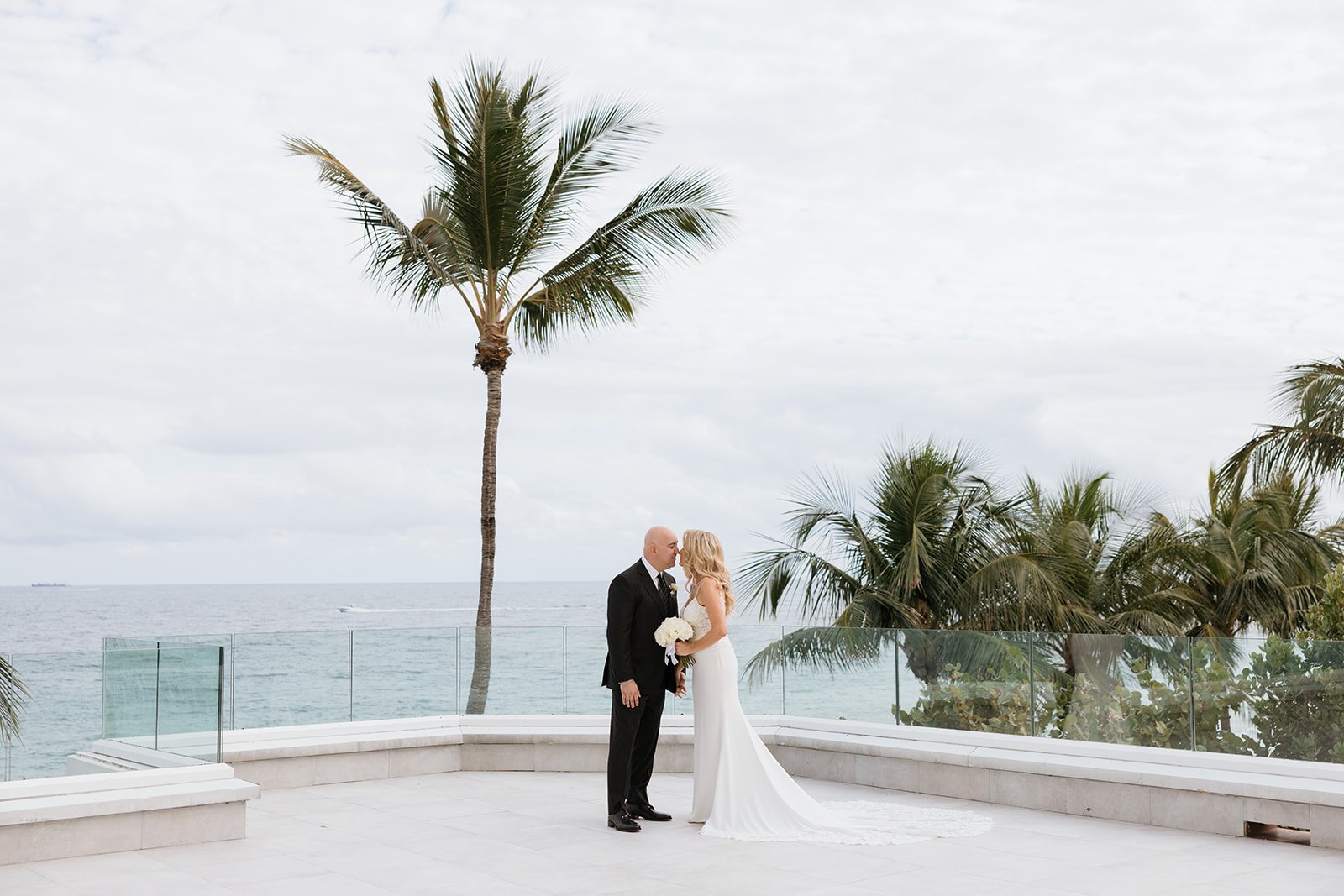 A bride and groom standing on a white tiled platform near the ocean, sharing a kiss with palm trees in the background.