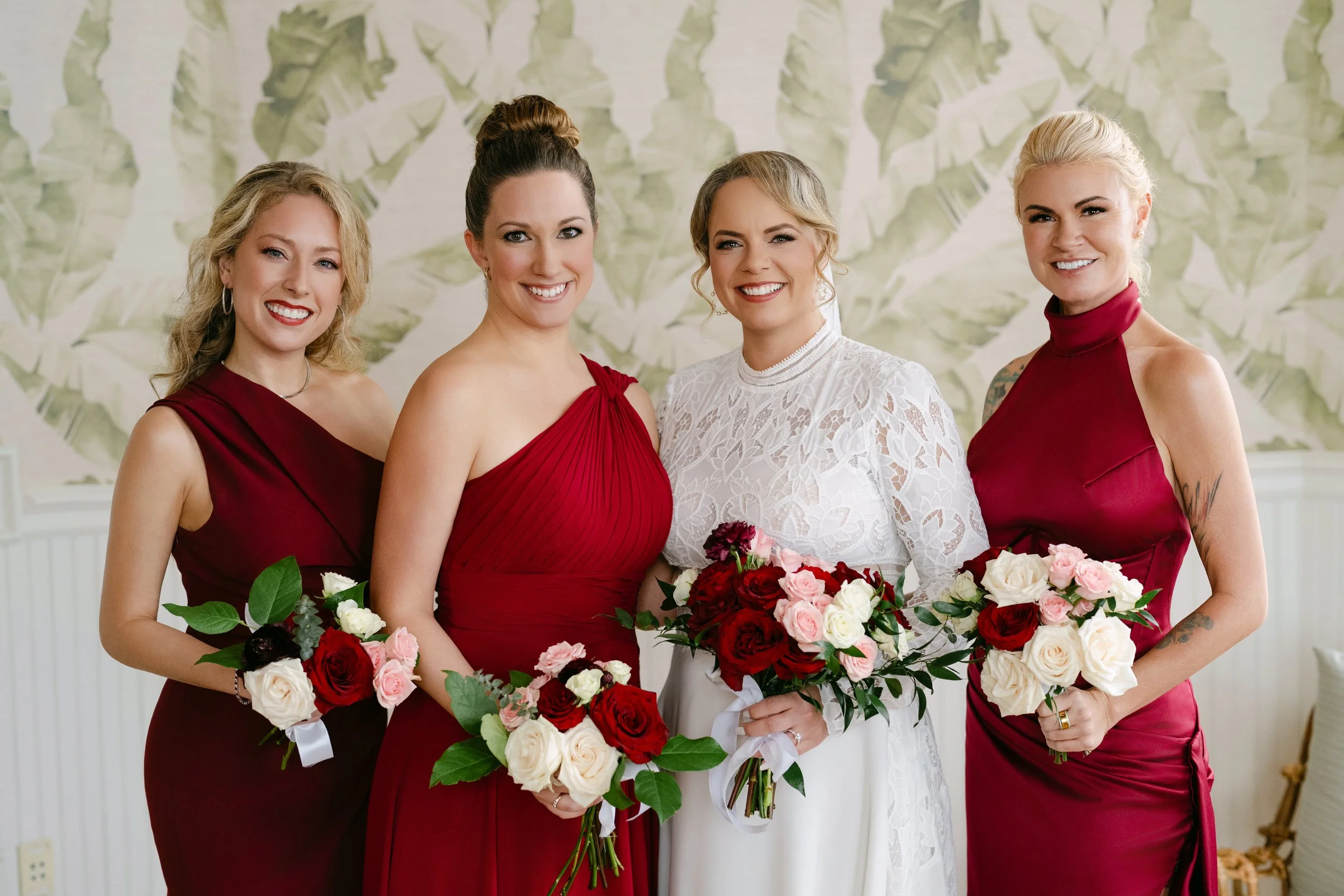 Four women in elegant dresses holding bouquets of flowers, standing together indoors, smiling at the camera.